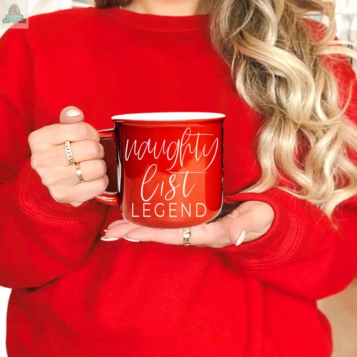 A person in a red sweater with long, wavy blonde hair holds the Naughty Legend Mug, a ceramic campfire mug with “naughty list LEGEND” in white script. Their festive look is complete with stylish rings and cozy holiday vibes.