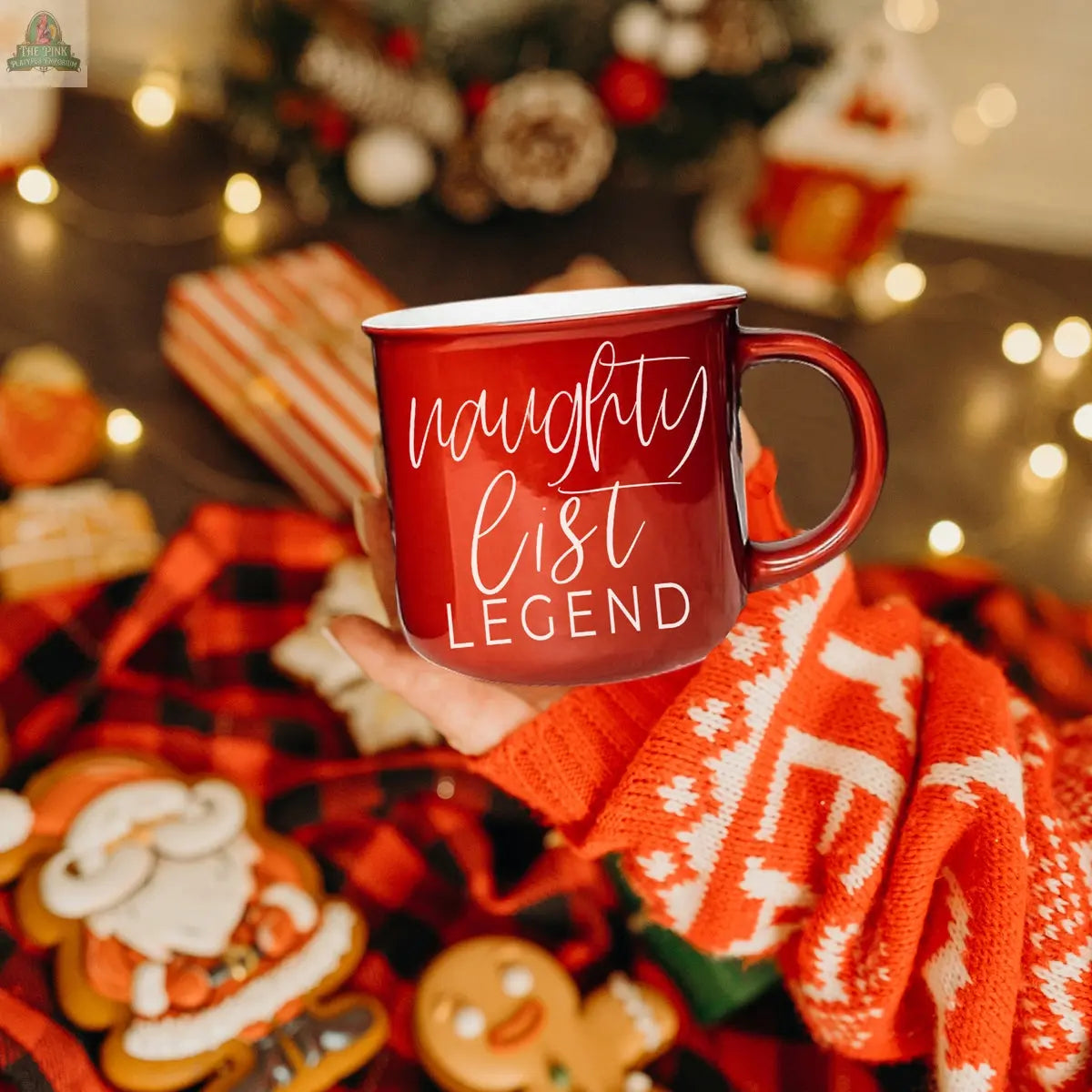 A hand in a red and white festive sweater holds the Naughty Legend Mug. Christmas cookies, wrapped gifts, and warm holiday lights in the background create the perfect festive setting for this holiday mug.