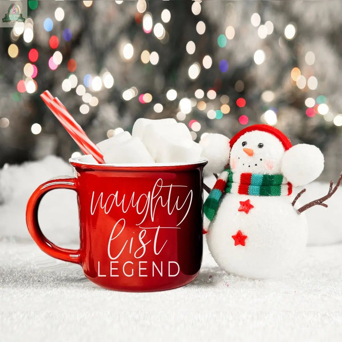 A red Naughty Legend Mug filled with marshmallows and candy canes sits beside a smiling snowman in a striped scarf, with festive holiday lights softly glowing in the background.