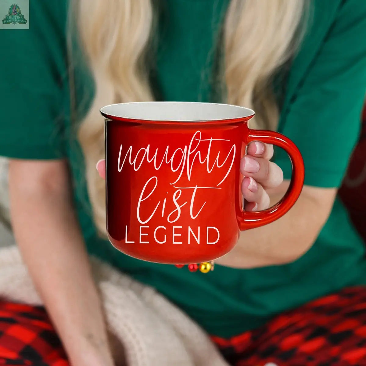 A person wearing a green shirt and red plaid pants holds the Naughty Legend Mug, a festive ceramic campfire mug with white "naughty list LEGEND" text.