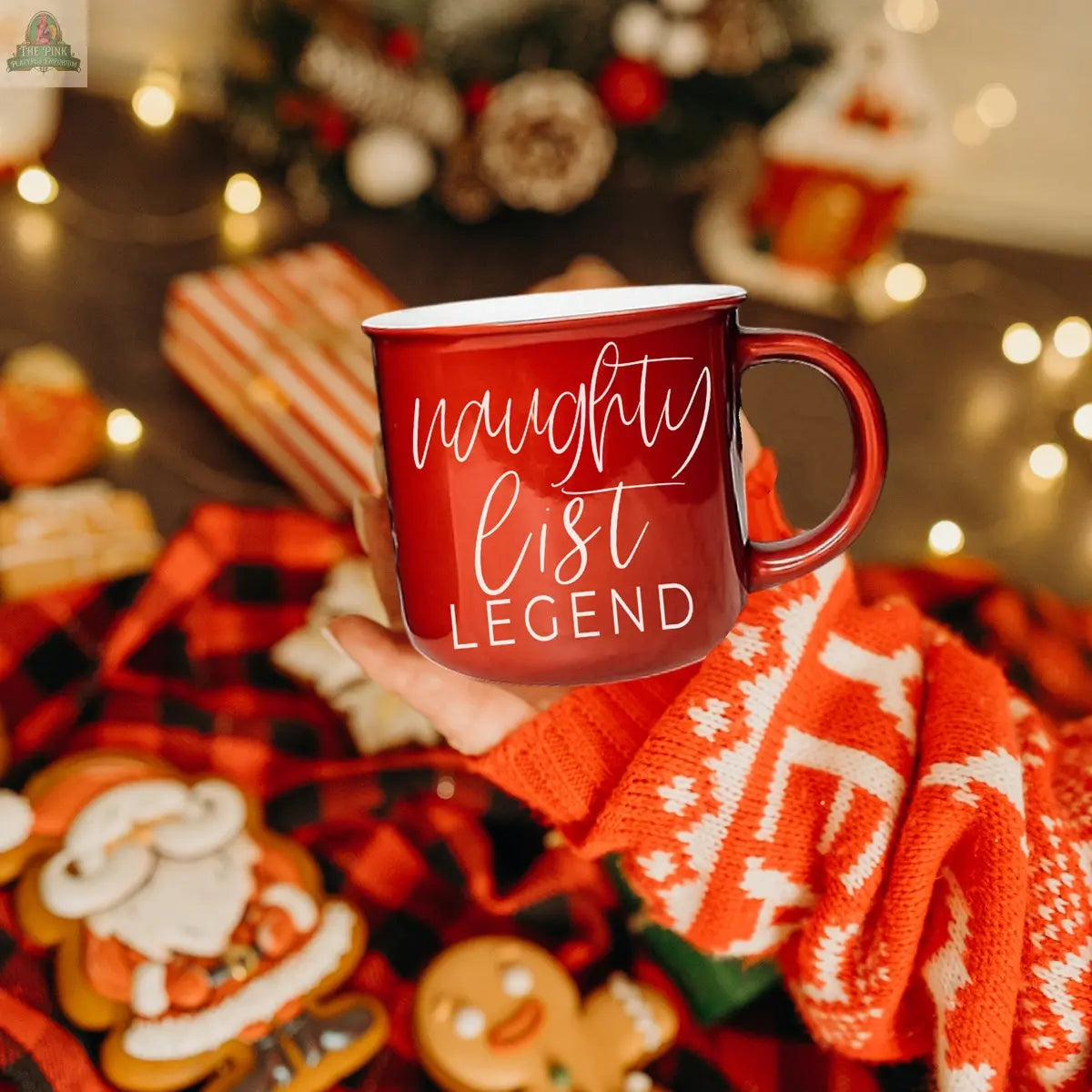 A hand in a festive red sweater holds the Naughty Legend Mug with “naughty list LEGEND.” Christmas cookies, wrapped gifts, and string lights in the background complete this cozy holiday scene.