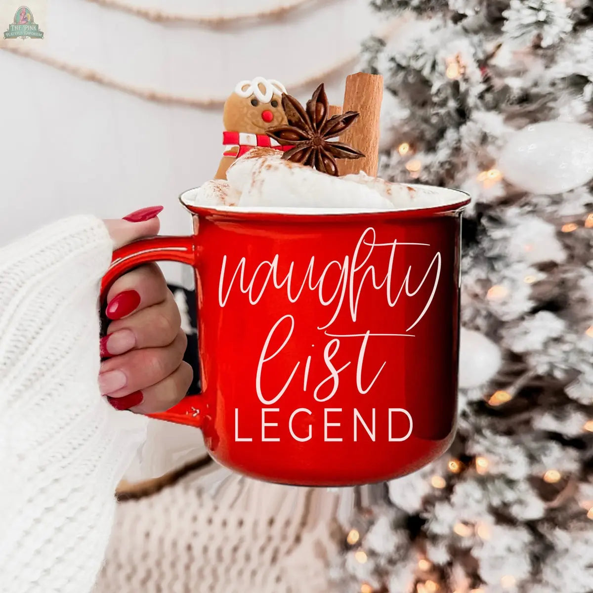 A hand with red nails holds a Naughty Legend Mug filled with whipped cream, topped with a gingerbread cookie, star anise, and a cinnamon stick. "naughty list LEGEND" is printed on the festive ceramic mug, with a snowy Christmas tree behind it.