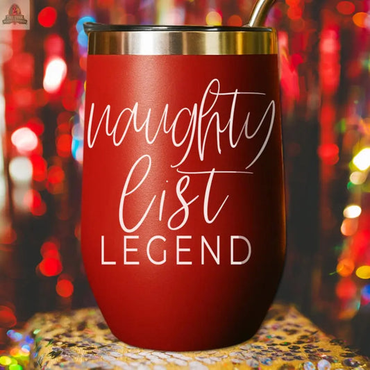 A red stainless steel Naughty List 17oz tumbler with a metal rim and straw sits on a festive surface. “naughty list LEGEND” is printed in white script, with colorful holiday lights creating a cheerful background.
