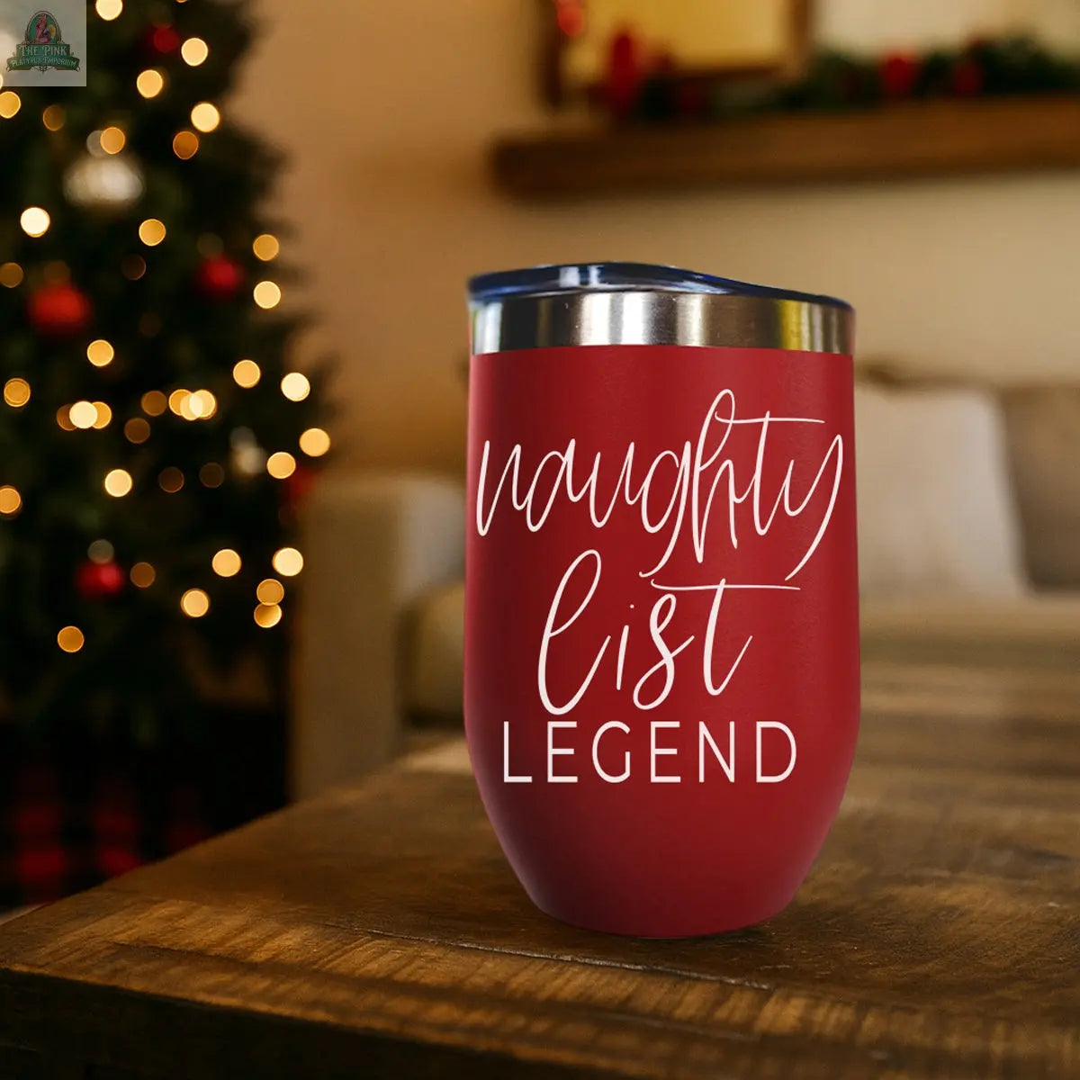 A Naughty List 17oz red stainless steel tumbler with "naughty list LEGEND" in white script rests on a wooden table, with a blurred Christmas tree and festive lights in the background.