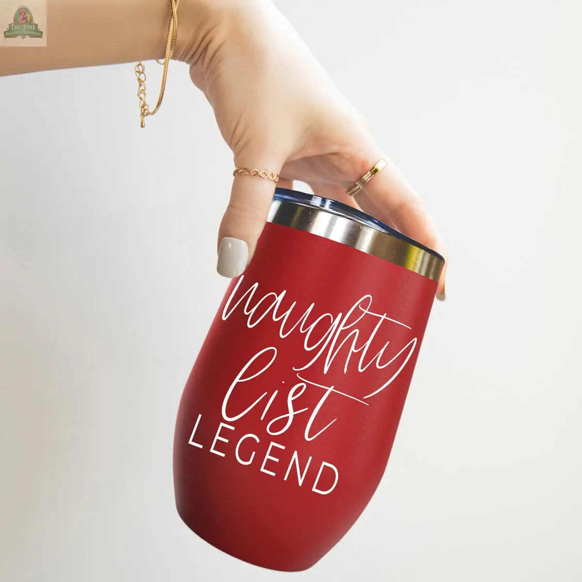 A hand with gold jewelry holds the Naughty List 17oz red stainless steel wine tumbler with a silver rim, featuring white handwritten-style text: naughty list LEGEND.