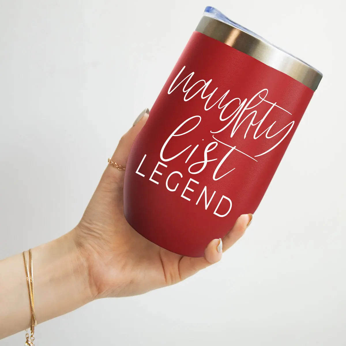 A hand holds the Naughty List 17oz red stainless steel tumbler with a silver rim, featuring "naughty list LEGEND" in white script and caps. The person also wears a bracelet and rings.