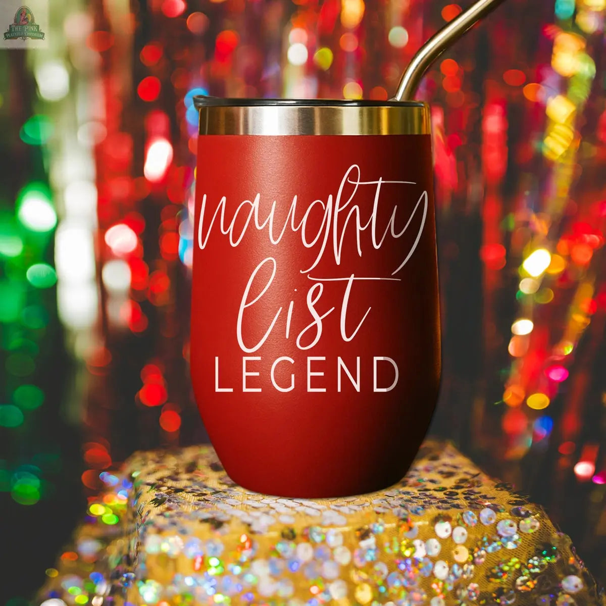A Naughty List 17oz red stainless steel tumbler with a metal straw rests on a glittery surface. “Naughty list LEGEND” in white script is printed on it. Colorful, blurred festive lights shine in the background.