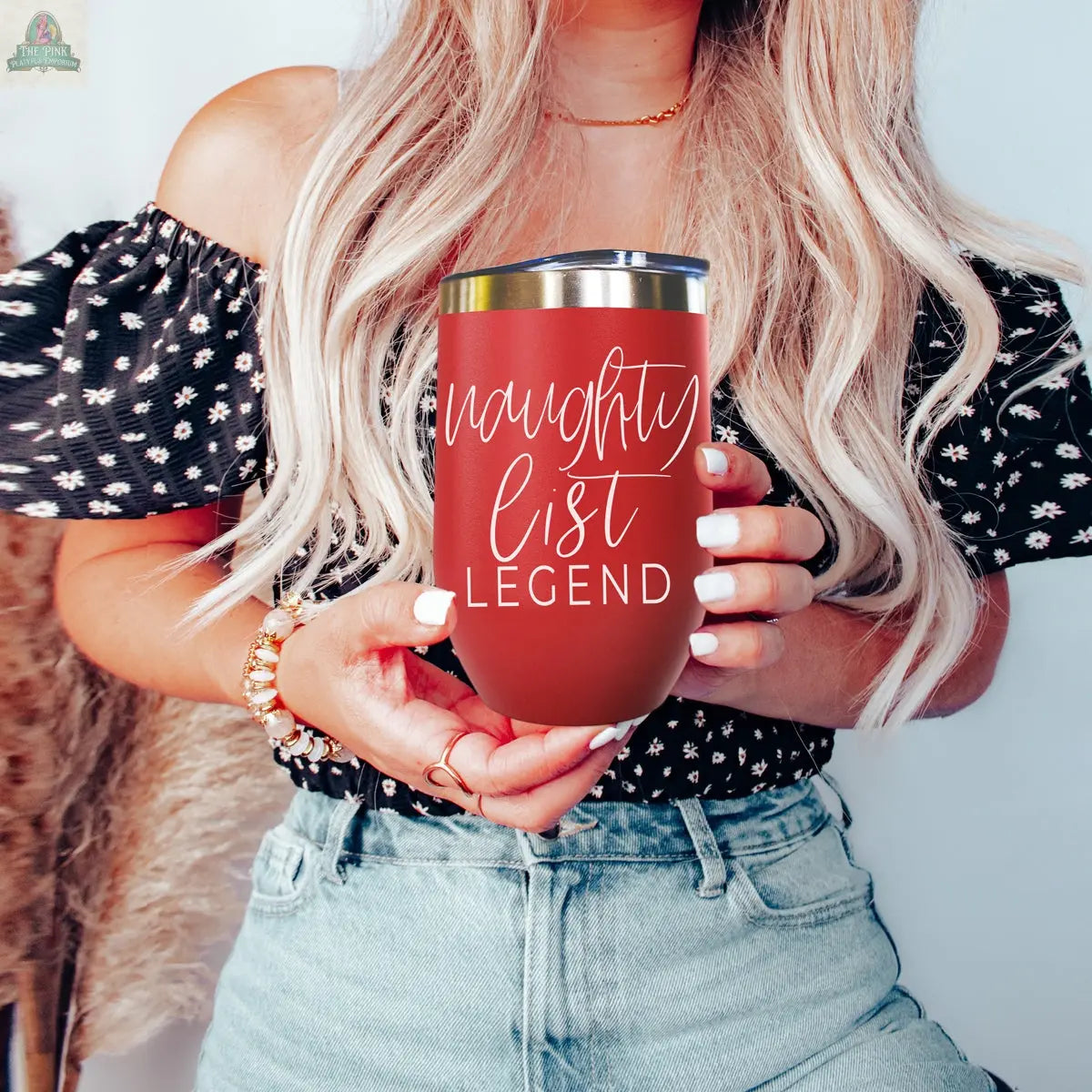 A woman with long blonde hair, in a black off-shoulder top and light blue jeans, holds the Naughty List 17oz tumbler—a red stainless steel cup with "naughty list LEGEND" in stylish white lettering.