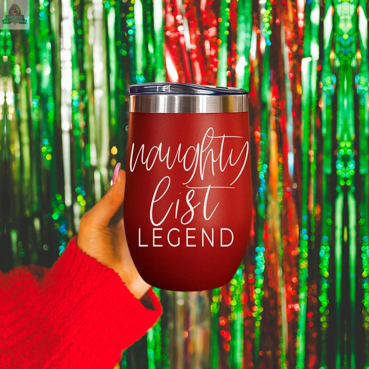 A hand in a red sweater holds the Naughty List 17oz stainless steel tumbler with “naughty list LEGEND” in white script, set against shimmering green and red tinsel.