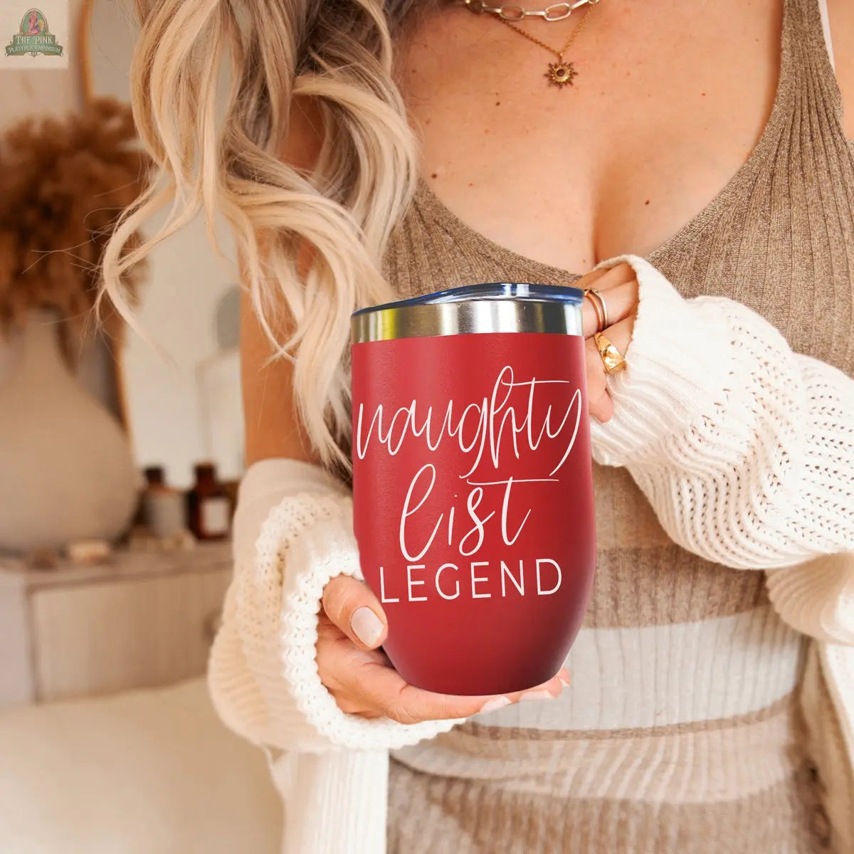 A woman with long blonde hair holds the Naughty List 17oz red stainless steel tumbler featuring “naughty list LEGEND” in white script. She wears a tan knit dress and white sweater, standing in a cozy, warmly lit room.
