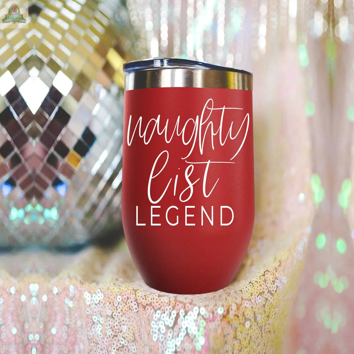 A red stainless steel Naughty List 17oz tumbler with a metal lid and "naughty list LEGEND" in white script is displayed on sequins. A disco ball in the background adds festive flair to this legendary wine glass alternative.