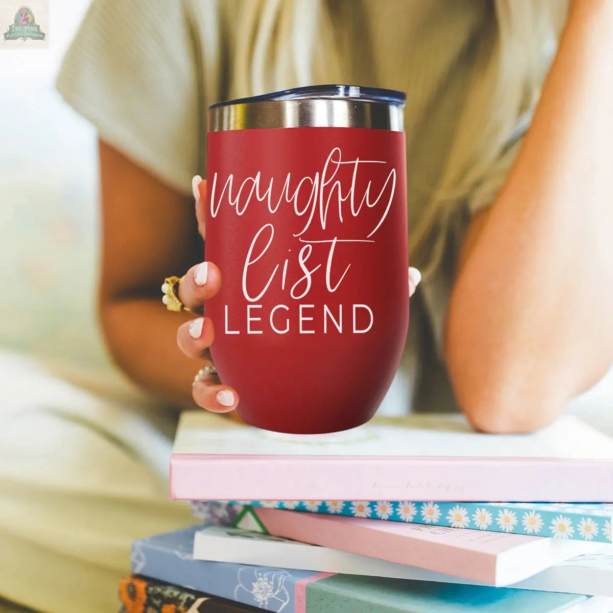 A person with long blonde hair holds the Naughty List 17oz red stainless steel tumbler with “naughty list legend” in white script, seated by colorful books. Only their torso and hand are visible.