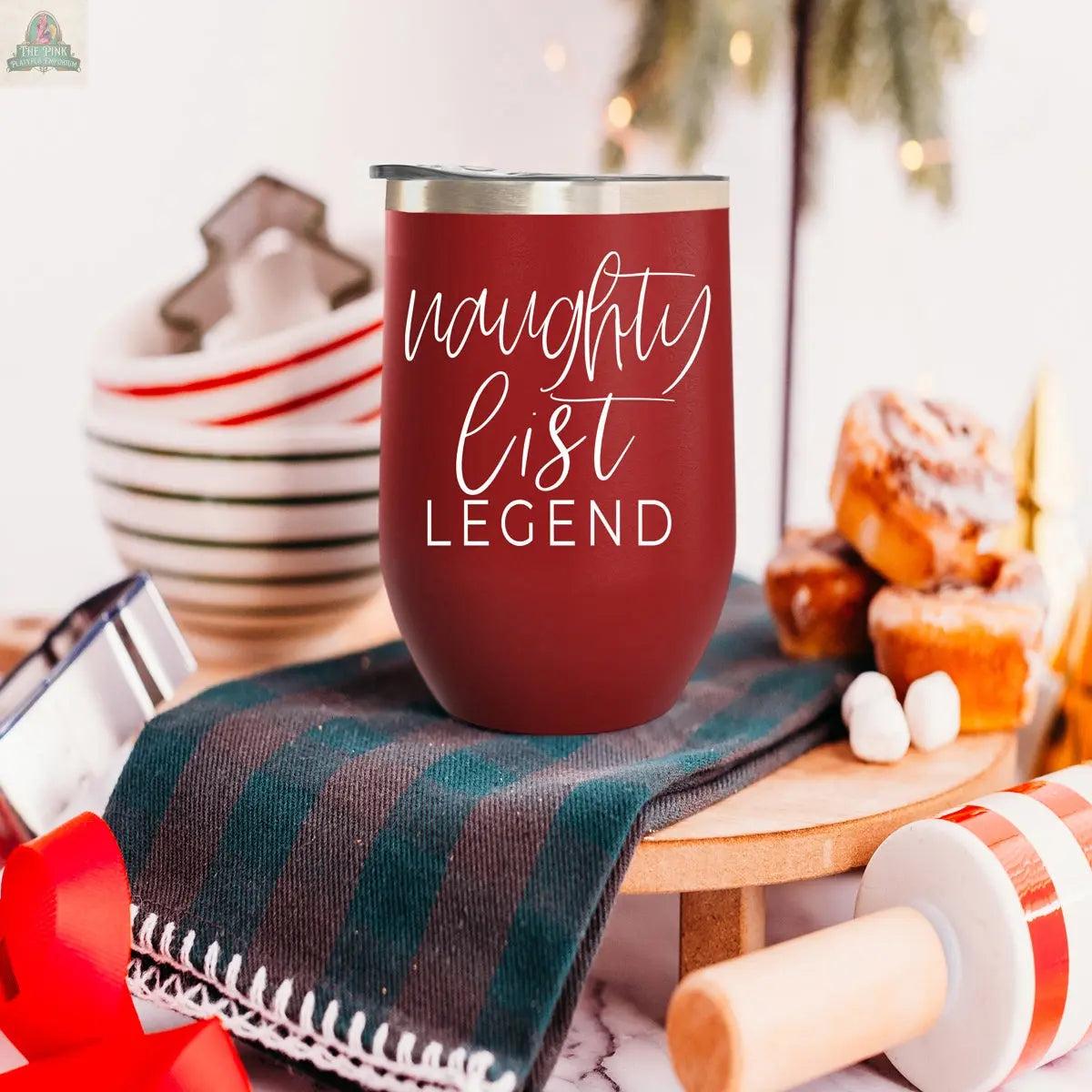 The Naughty List 17oz maroon stainless steel tumbler, featuring "NAUGHTY LIST LEGEND," rests on a striped cloth amid holiday treats, plates, and festive decorations.