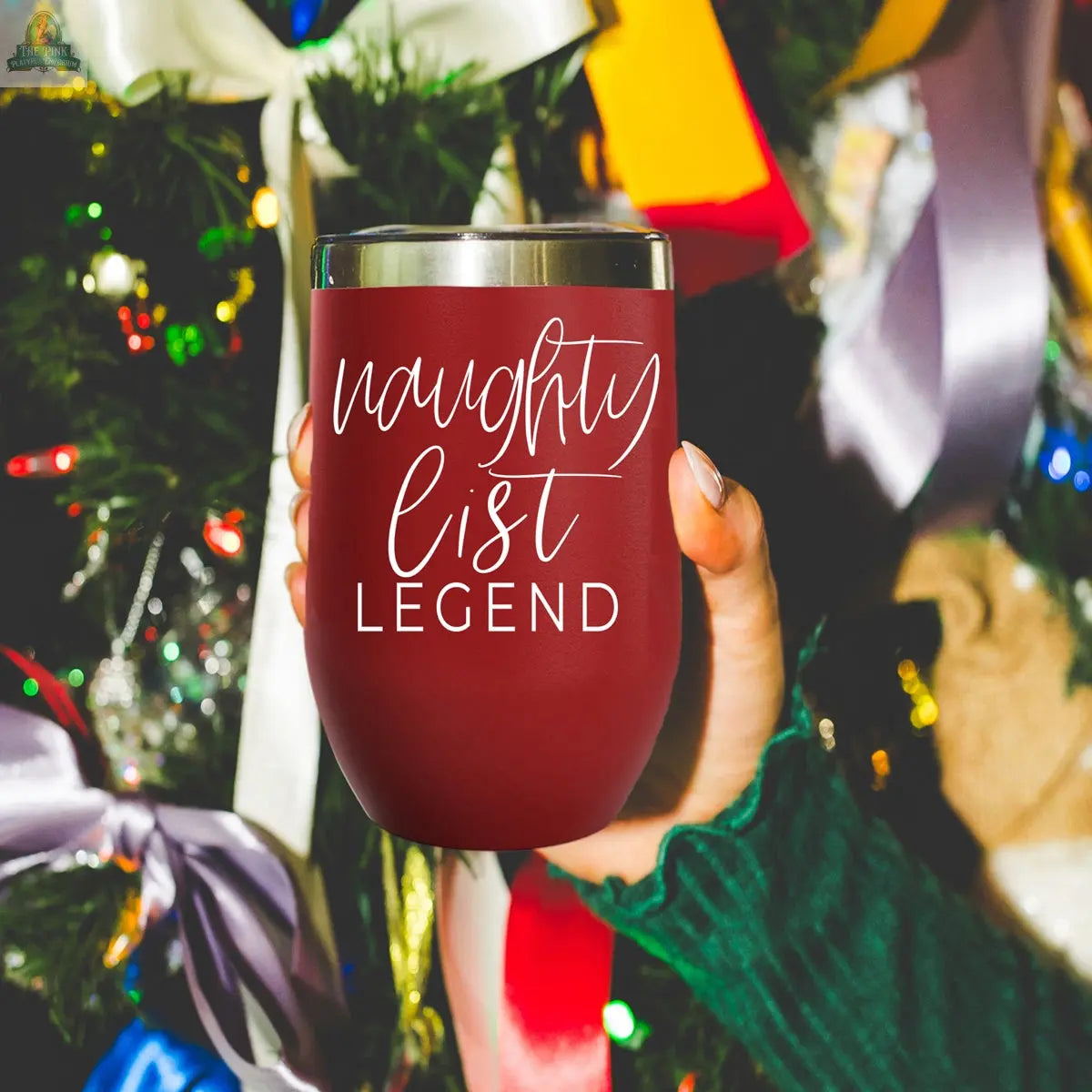 A hand in a green sweater holds the Naughty List 17oz red stainless steel tumbler with "naughty list LEGEND" printed on it, in front of a decorated Christmas tree with colorful lights and ribbons.