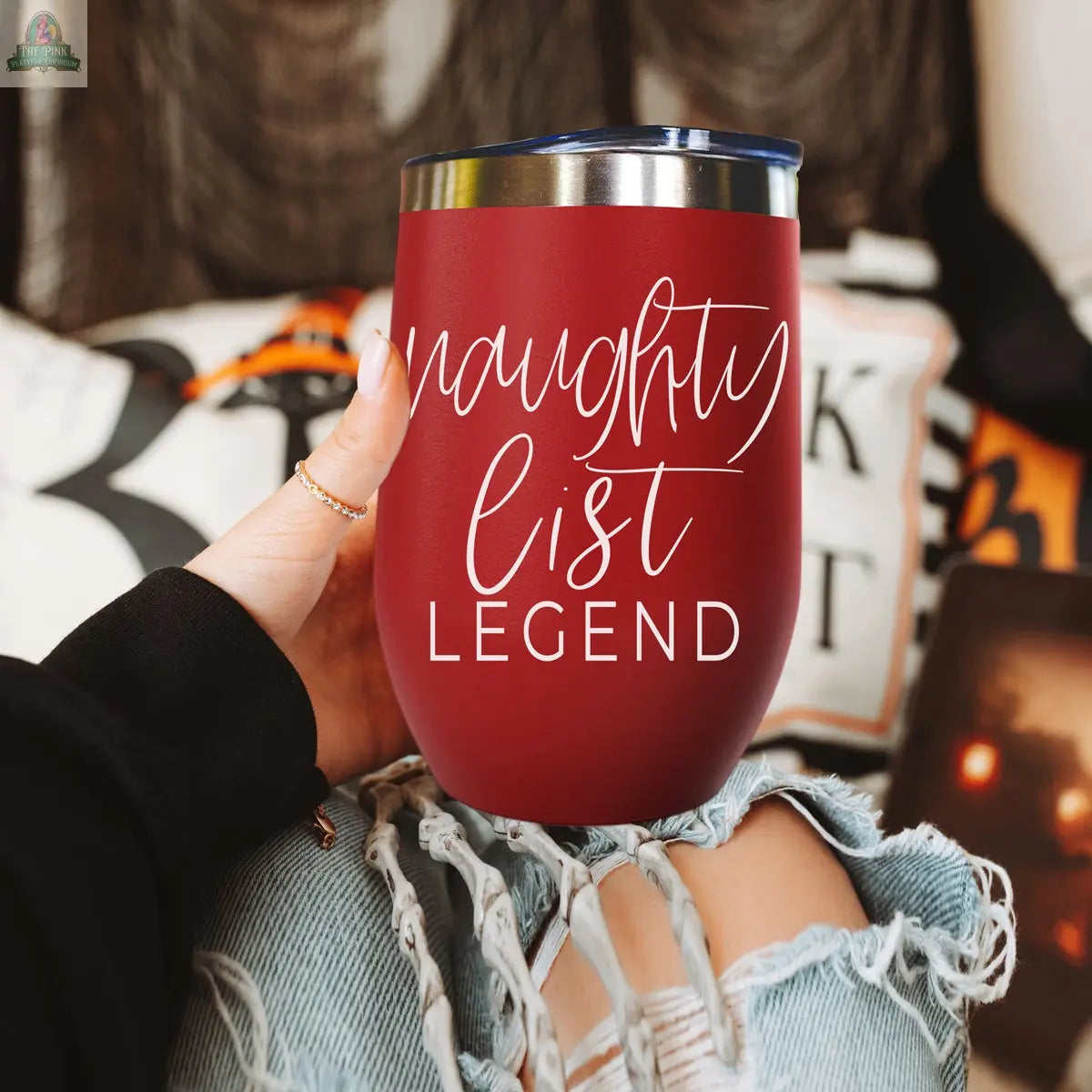 A person with ripped jeans holds a red Naughty List 17oz stainless steel tumbler featuring “naughty list legend” in white script, surrounded by cozy pillows and blankets in the background.