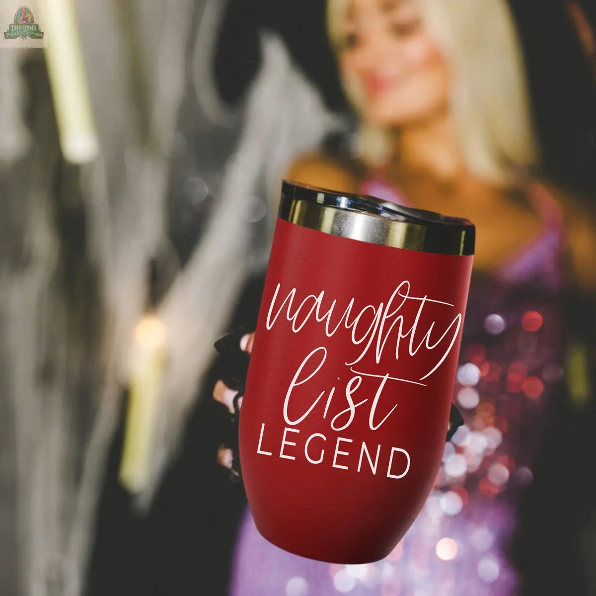 A woman in a sparkly purple dress holds up the Naughty List 17oz red stainless steel tumbler with "naughty list LEGEND" in white script. The blurred background features faint lights and visible Halloween decorations.