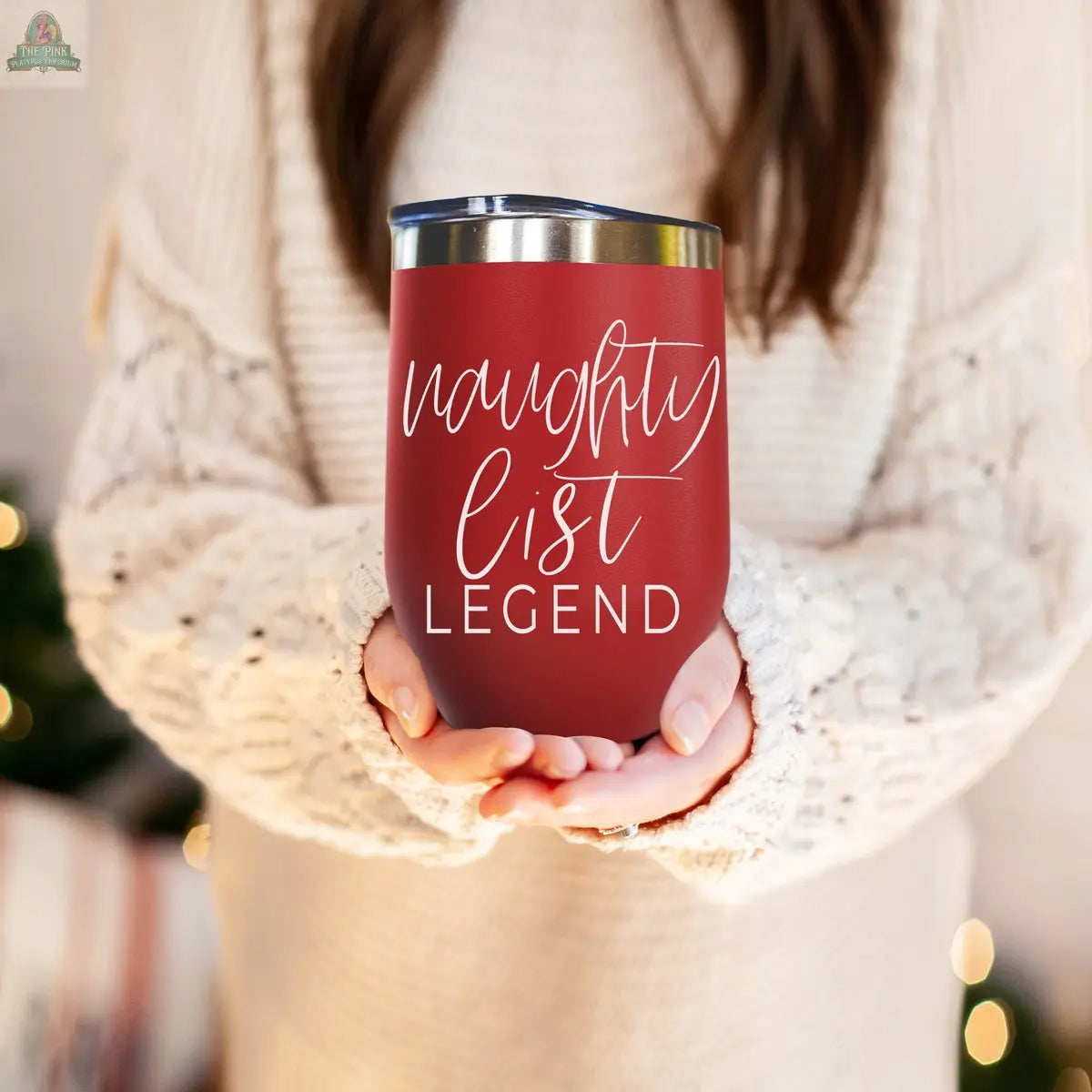 A person in a cream knit sweater holds the Naughty List 17oz red wine tumbler, featuring "naughty list LEGEND" written in white script.