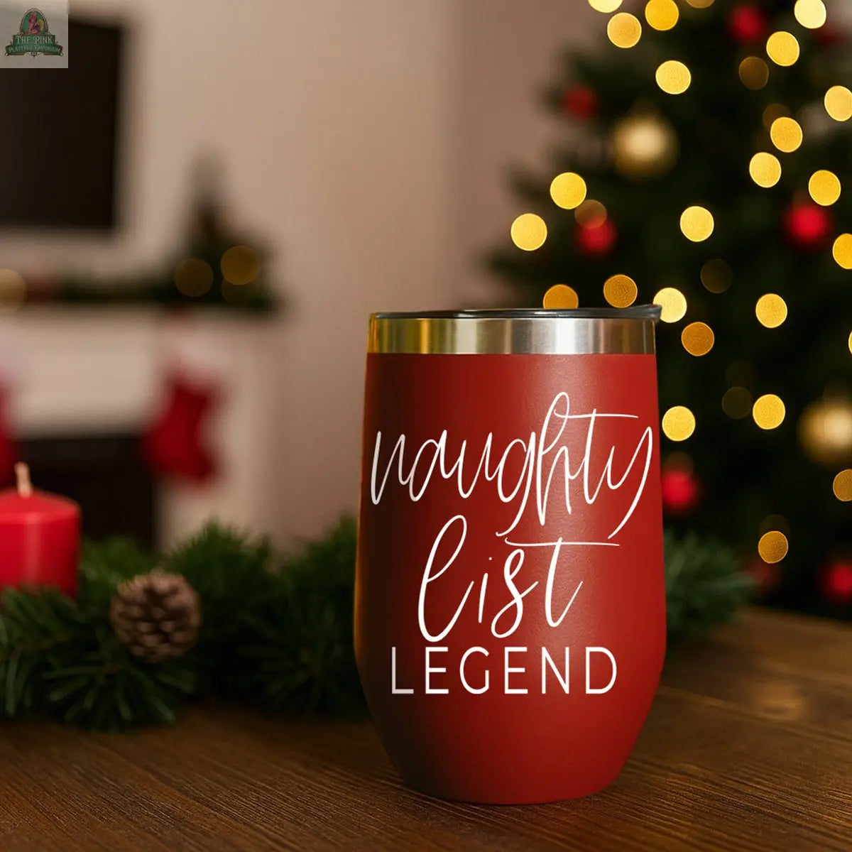 A Naughty List 17oz red stainless steel tumbler with "naughty list LEGEND" in white sits on a wooden table among Christmas decorations, candles, and a blurred lit Christmas tree in the background.