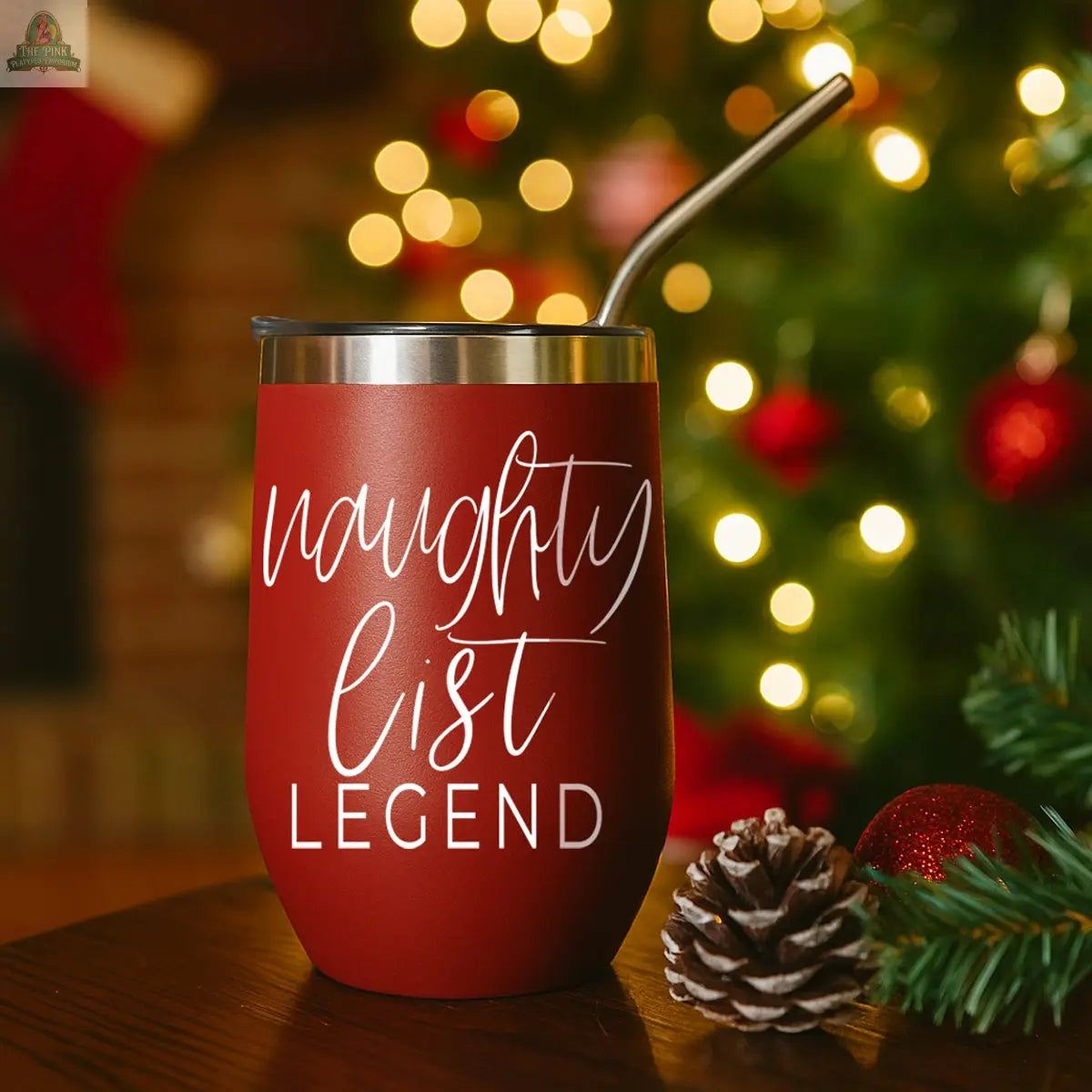 The Naughty List 17oz red stainless steel tumbler with a metal straw features “naughty list LEGEND” in white script. It's displayed near a pine cone, glowing Christmas tree, and a stocking by the fireplace.