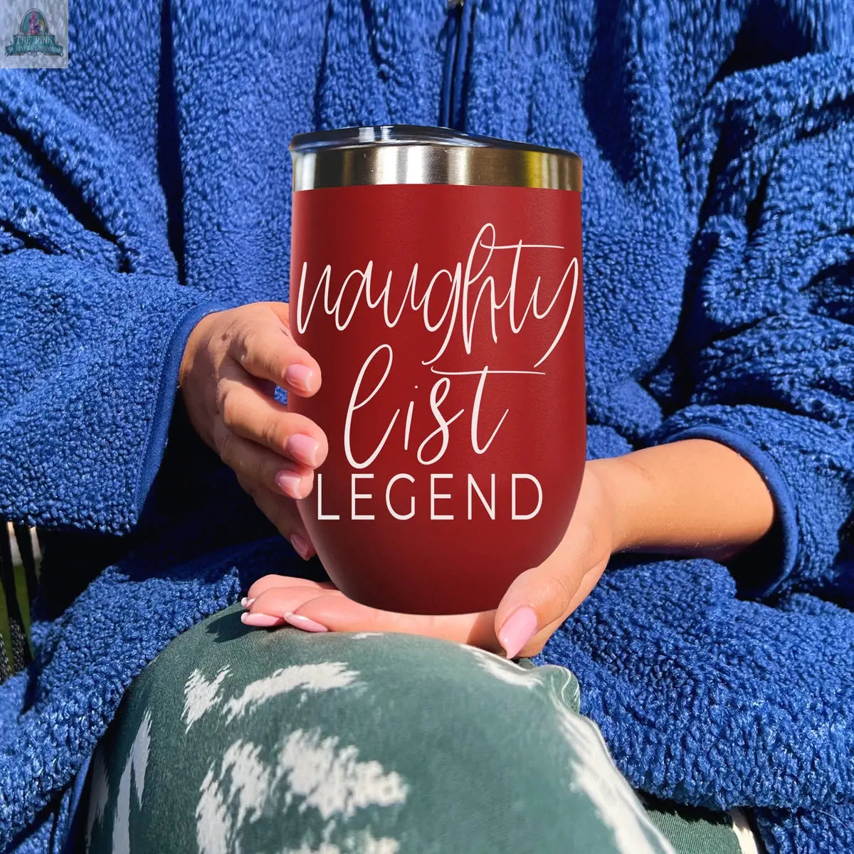A person in a blue fleece jacket and green patterned pants holds a red Naughty List 17oz stainless steel tumbler with "naughty list LEGEND" in white script.