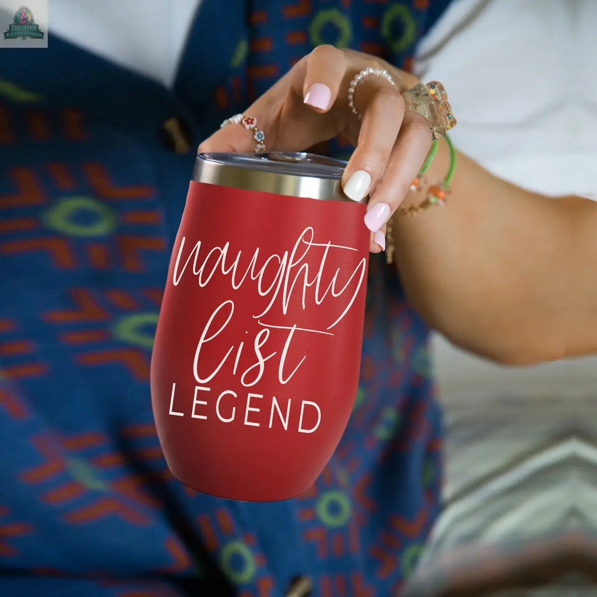 A person holds the Naughty List 17oz red stainless steel tumbler with "naughty list LEGEND" in white script. Their manicured nails, rings, and bracelets contrast with a patterned blue and red garment in the background.