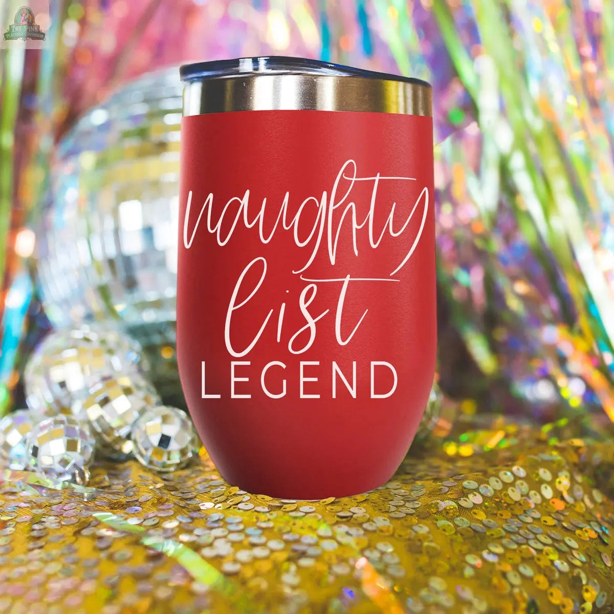 The Naughty List 17oz red stainless steel tumbler, featuring "naughty list LEGEND" in white script, sits on gold sequins with disco balls and vibrant streamers in the background.