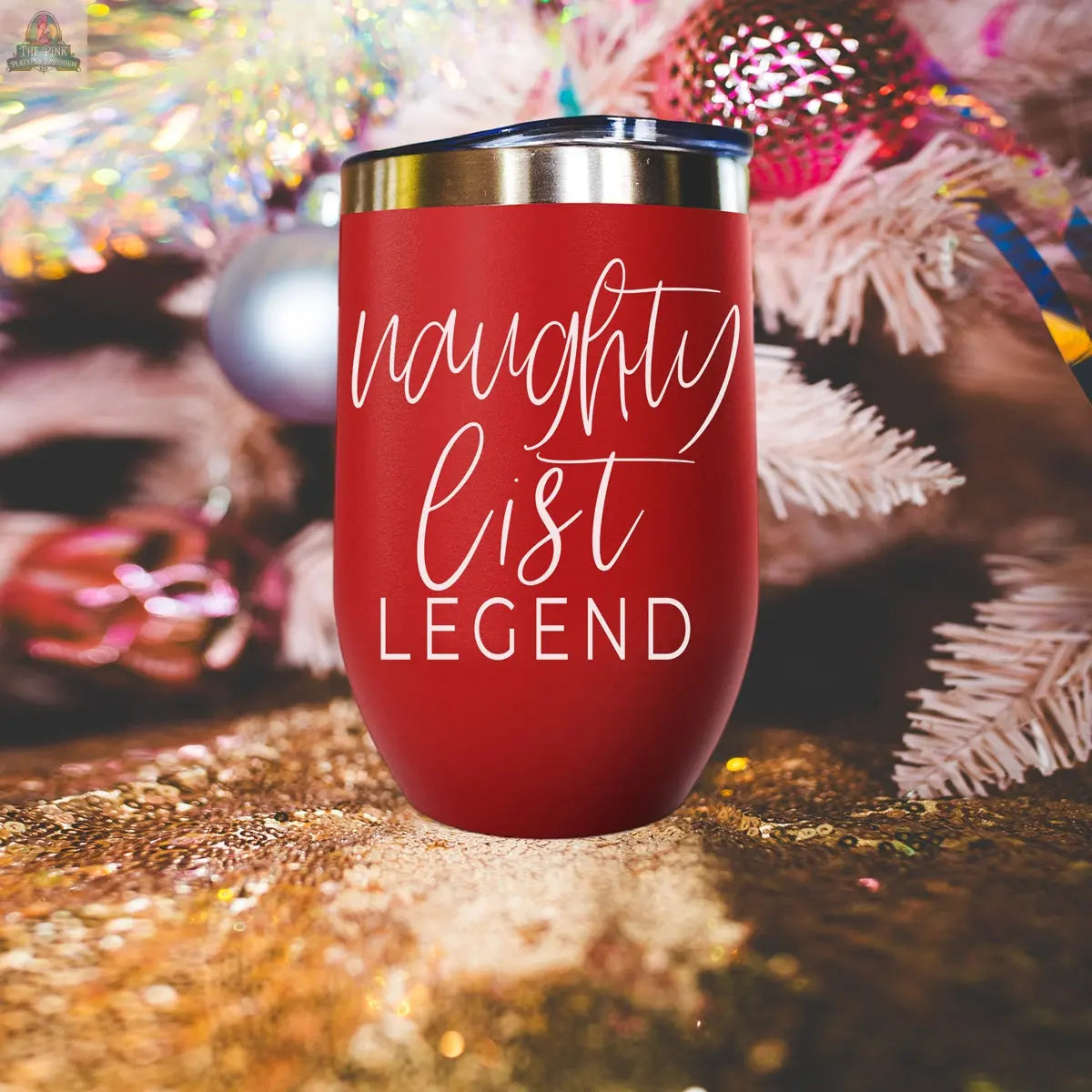 The Naughty List 17oz red stainless steel tumbler, featuring “naughty list LEGEND,” stands on a glittery gold surface with a blurred white Christmas tree and ornaments in the background.