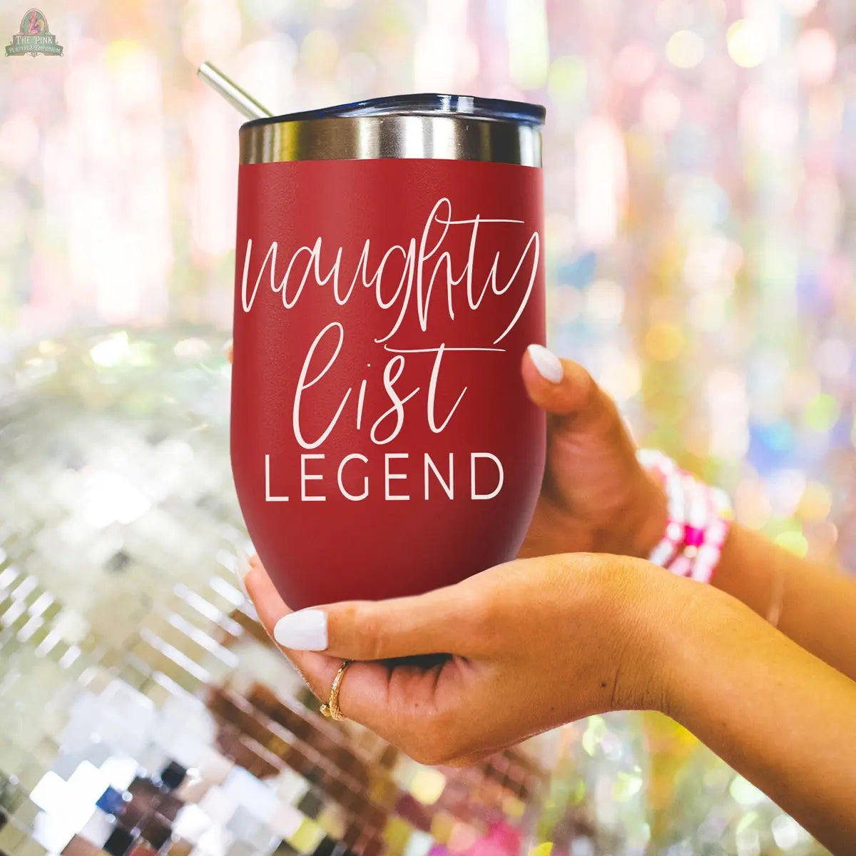 A person holds the Naughty List 17oz red stainless steel tumbler with a metal straw. “Naughty list LEGEND” is written in white script. A disco ball, vibrant sparkly background, and beaded bracelets are also visible.