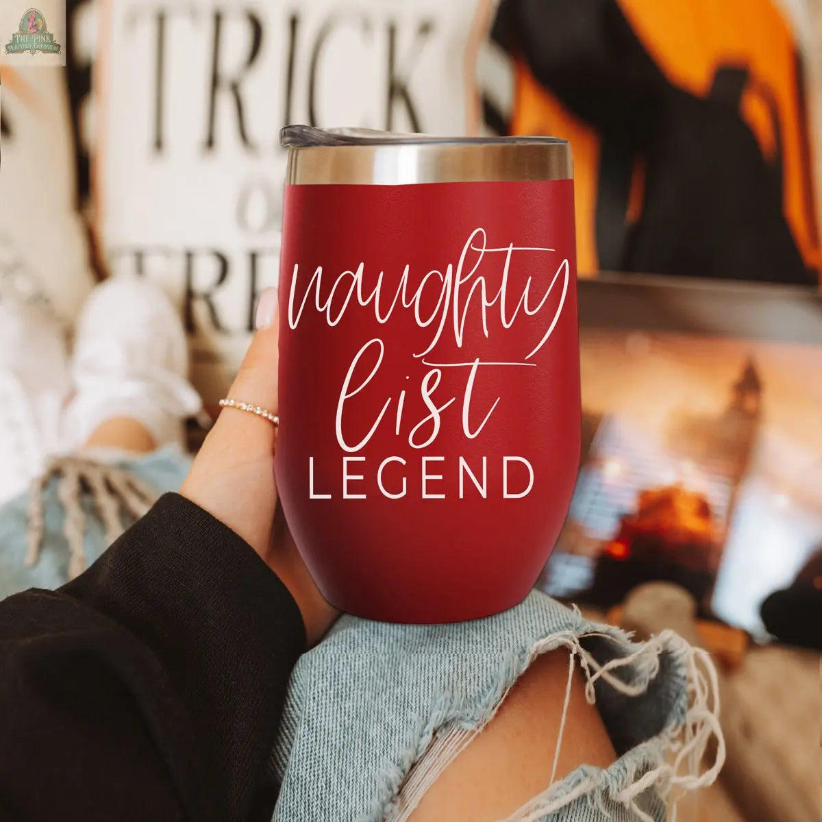 A person in ripped jeans holds a red Naughty List 17oz tumbler with "naughty list legend" in white script. In the blurred background, a pillow with "TRICK" and a laptop screen can be seen.