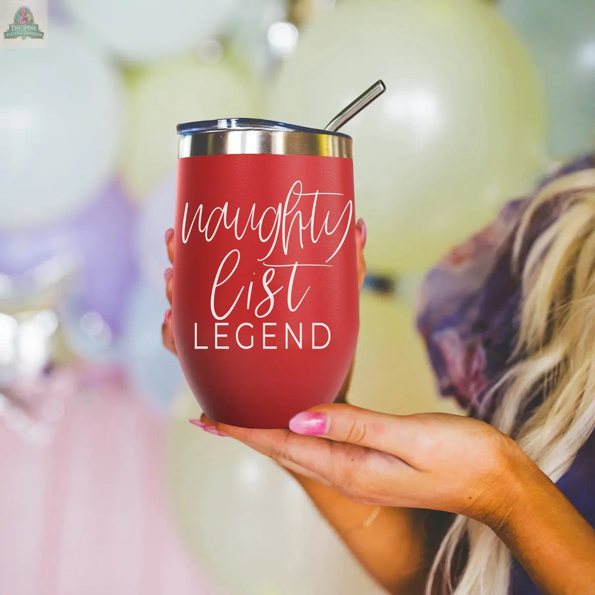 A person with long blonde hair holds a red Naughty List 17oz stainless steel tumbler with a metal straw and white script. Colorful balloons are blurred in the background.