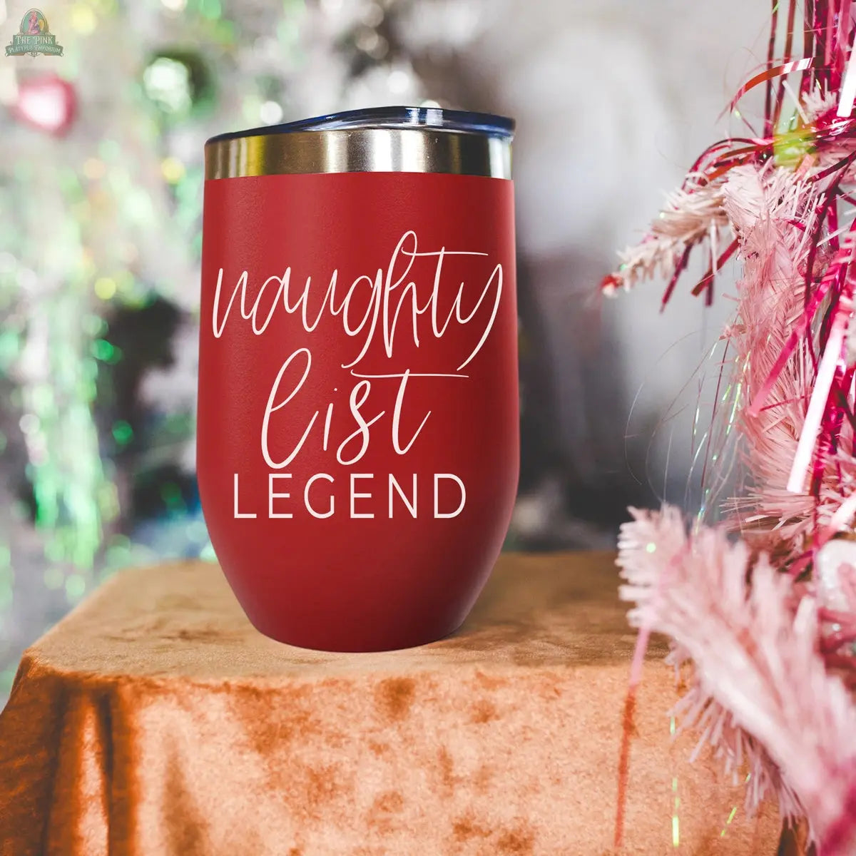 A Naughty List 17oz red stainless steel tumbler with a silver rim rests on a brown surface, featuring white script text reading "naughty list LEGEND." Blurred pink and green holiday decorations add a festive backdrop. Perfect for holiday fun.