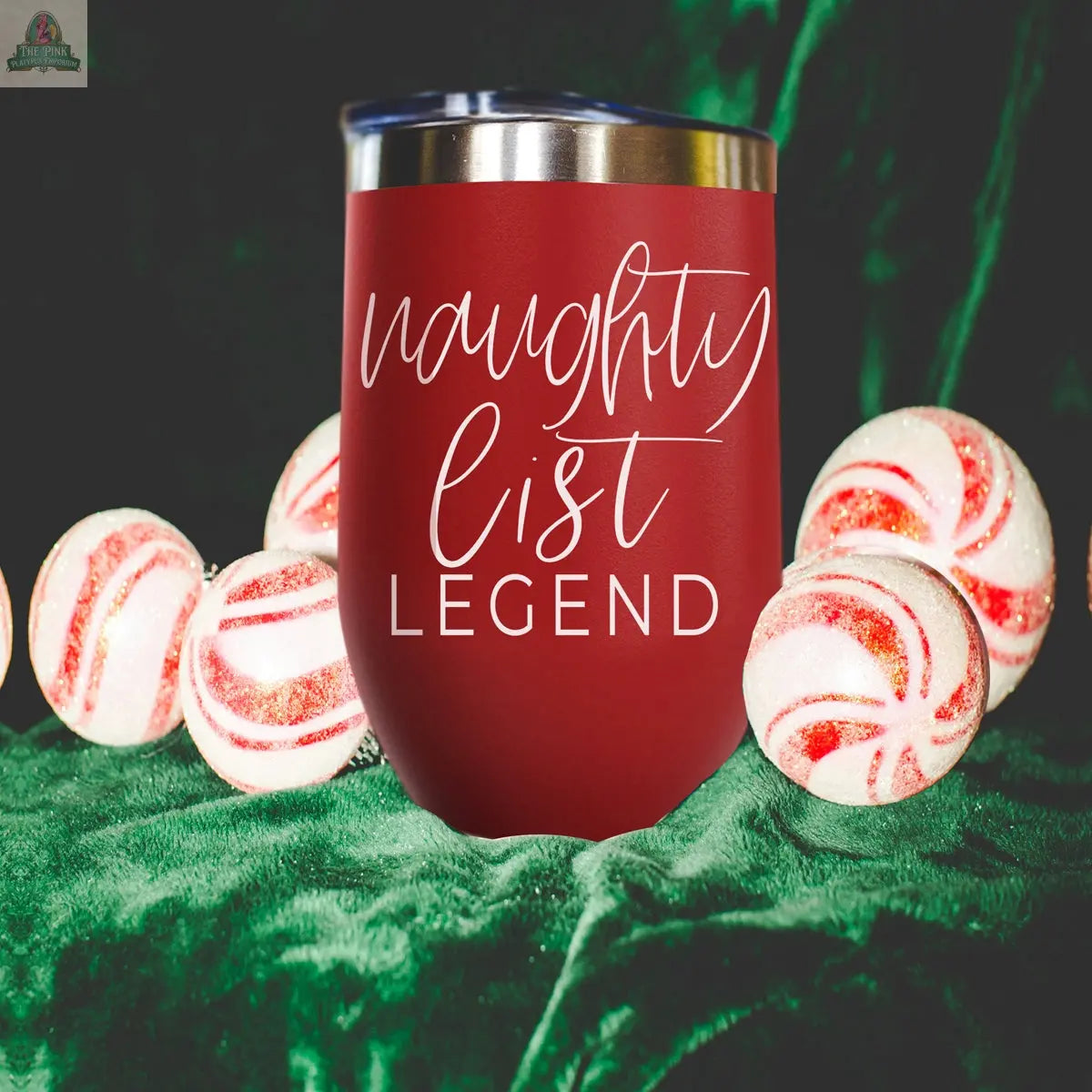 The Naughty List 17oz red stainless steel tumbler, featuring “naughty list LEGEND” in white script, rests on green velvet and is surrounded by red and white striped peppermint candies.