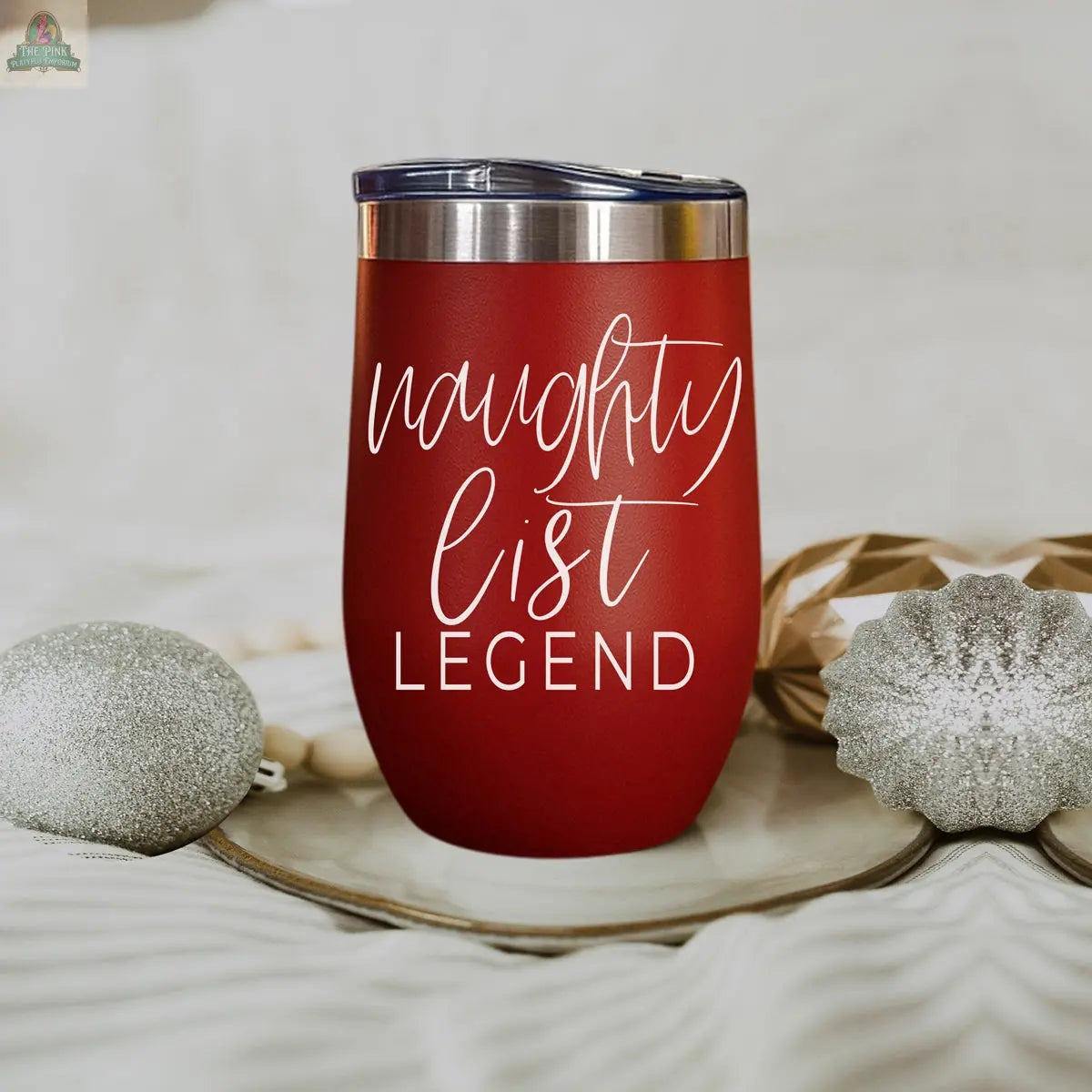 A red stainless steel Naughty List 17oz tumbler with a clear lid sits on a plate, surrounded by silver Christmas ornaments. White text on the tumbler reads “naughty list LEGEND.”.