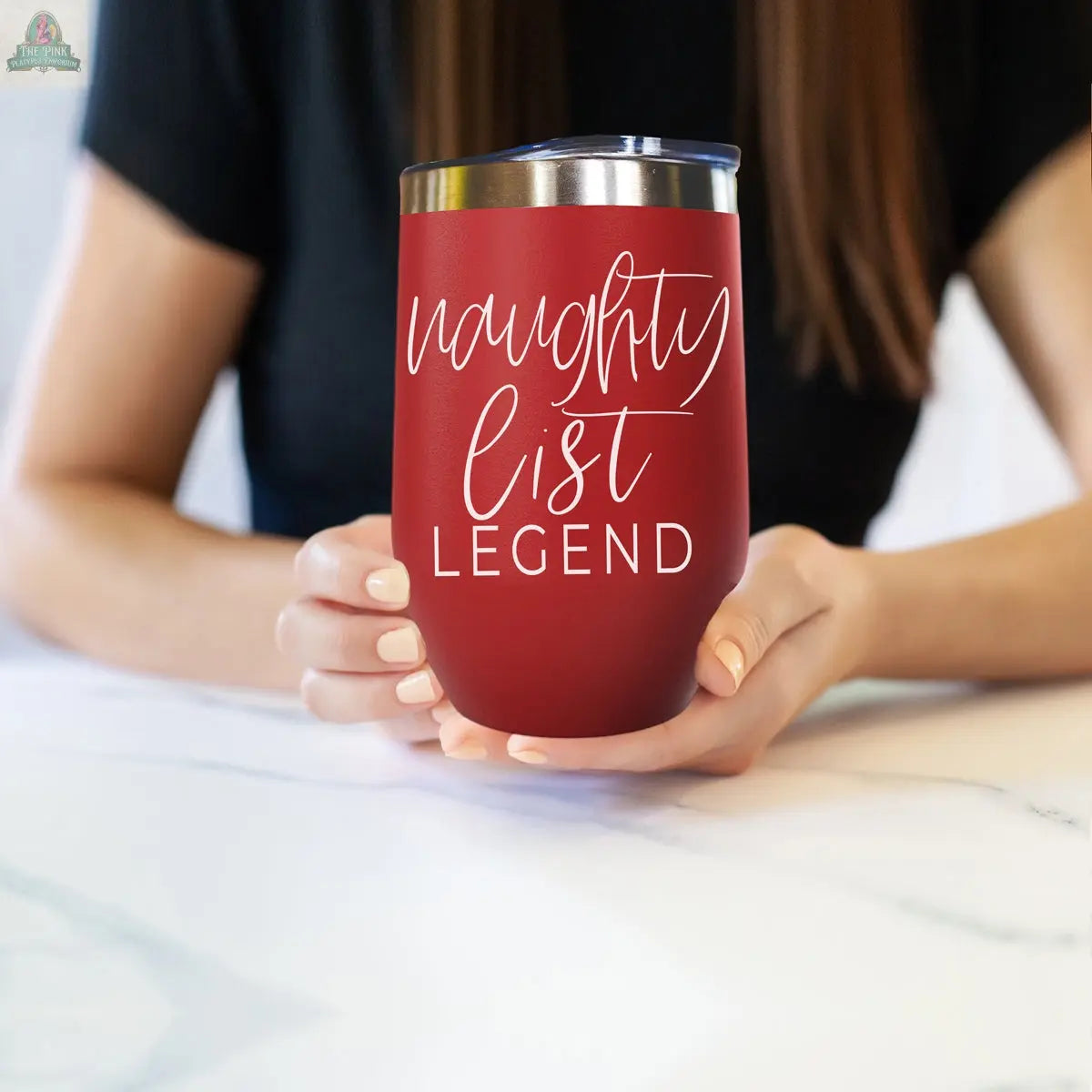 A person with long brown hair holds the Naughty List 17oz tumbler, featuring white "naughty list LEGEND" text. The blurred background draws focus to the tumbler and their hands.