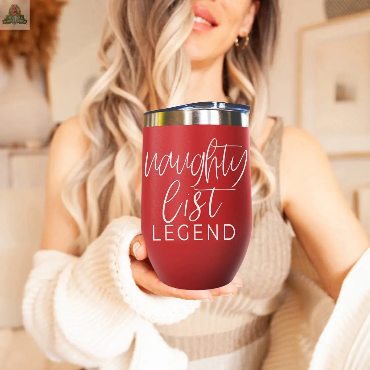 Smiling in a cozy, softly lit room, a woman with wavy hair holds the Naughty List 17oz red stainless steel tumbler, featuring "naughty list LEGEND" in white script. She wears a cream-colored sweater.