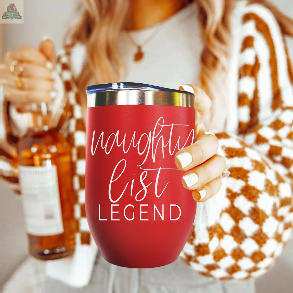 A person wearing a checkered sweater holds the Naughty List 17oz red stainless steel tumbler with "naughty list LEGEND" in white script, while holding a bottle in the other hand.
