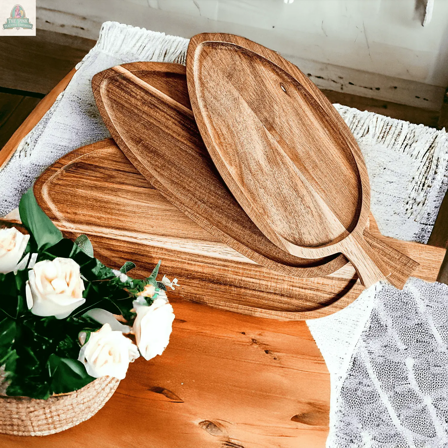 The Nesting Trays | 3 Sizes, made of oval acacia wood with handles, are stacked on a wooden table beside a small basket of white roses and a partially covering textured white cloth.