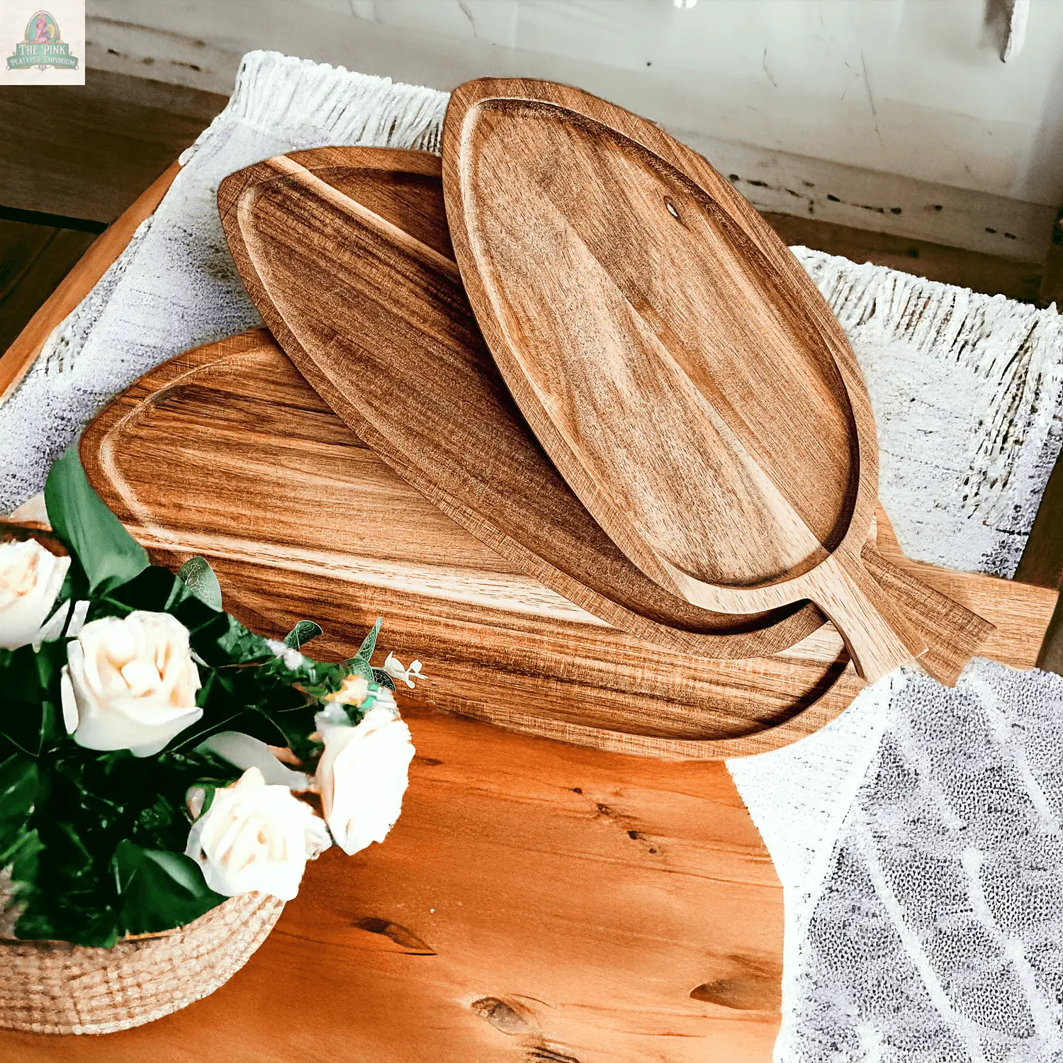 The Nesting Trays | 3 Sizes, made of oval acacia wood with handles, are stacked on a wooden table beside a small basket of white roses and a partially covering textured white cloth.