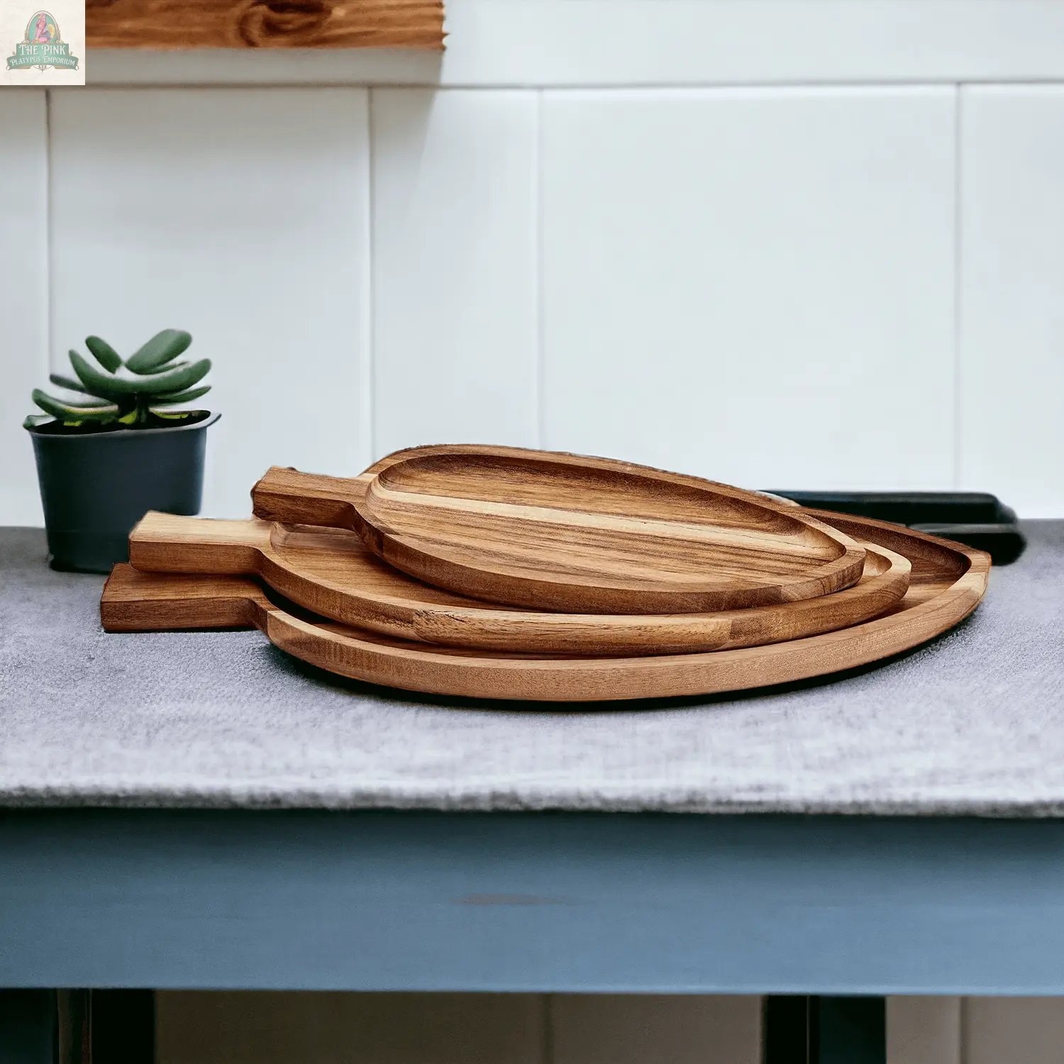 Three Nesting Trays | 3 Sizes, made of acacia wood with handles, are stacked on a gray cloth atop the counter. In the background, a small potted succulent sits by a white tiled wall.