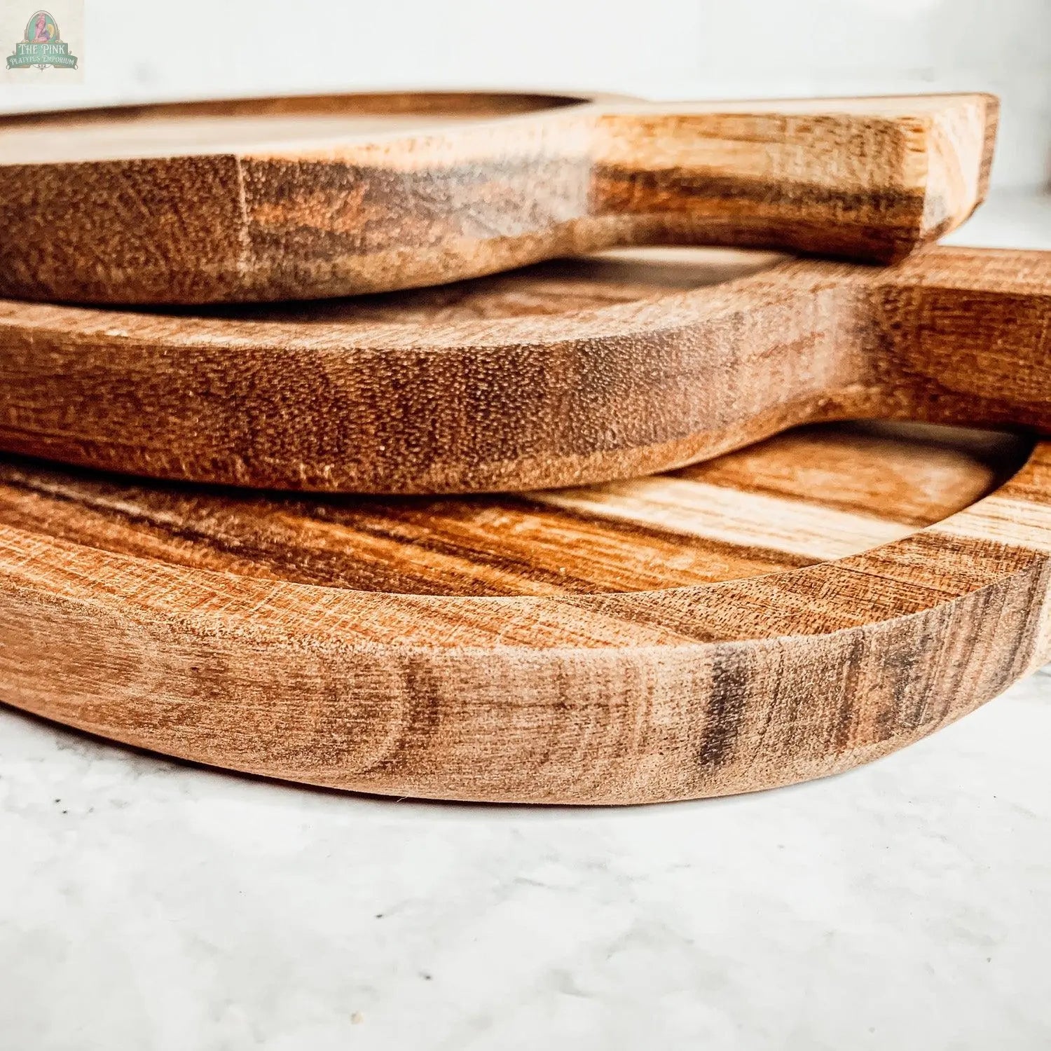 Three Nesting Trays in acacia wood are stacked on a white marble countertop, highlighting their handles, smooth rounded edges, and beautiful grain patterns.