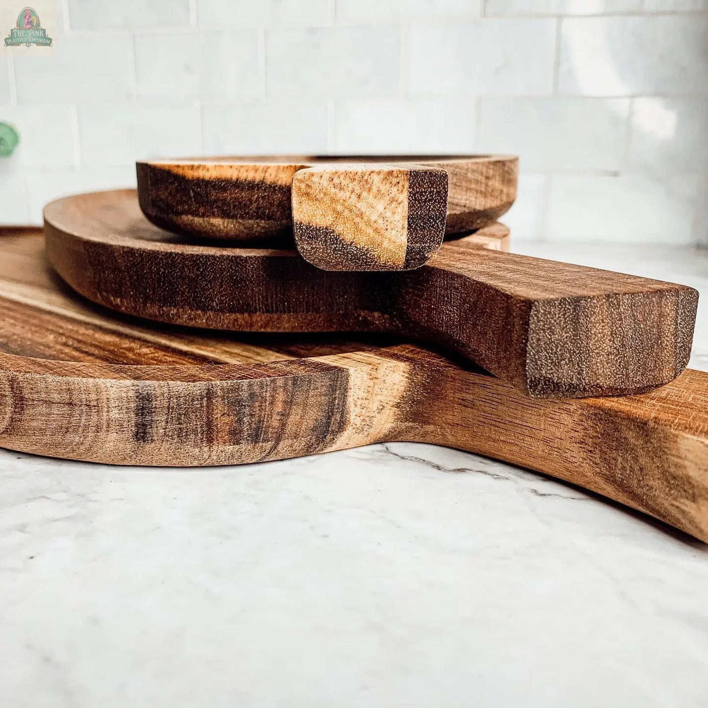 Three Nesting Trays | 3 Sizes, crafted from acacia wood and inspired by classic cheese boards, are stacked with aligned handles on a marble countertop, set against a white tiled backsplash.