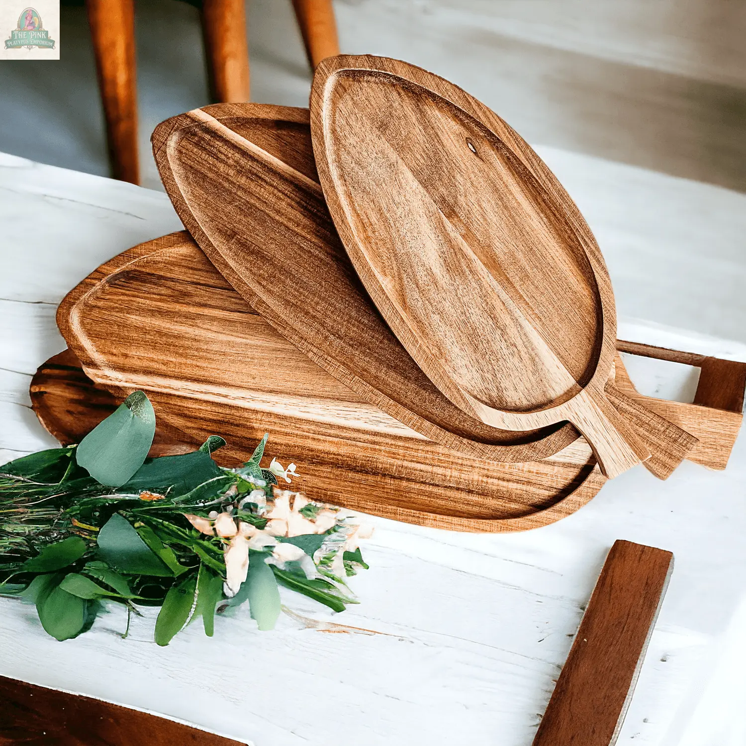 The Nesting Trays | 3 Sizes set features three acacia wood serving trays in unique shapes, stacked on a white table beside a leafy branch—perfect as cheese boards or leaf-shaped platters for stylish entertaining.