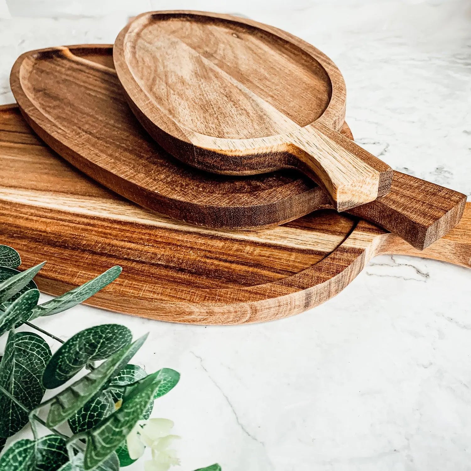 Three Nesting Trays in acacia wood with handles are stacked on a white marble surface, highlighting their natural wood grain. A green leafy plant peeks in the corner. Ideal as cheese boards.