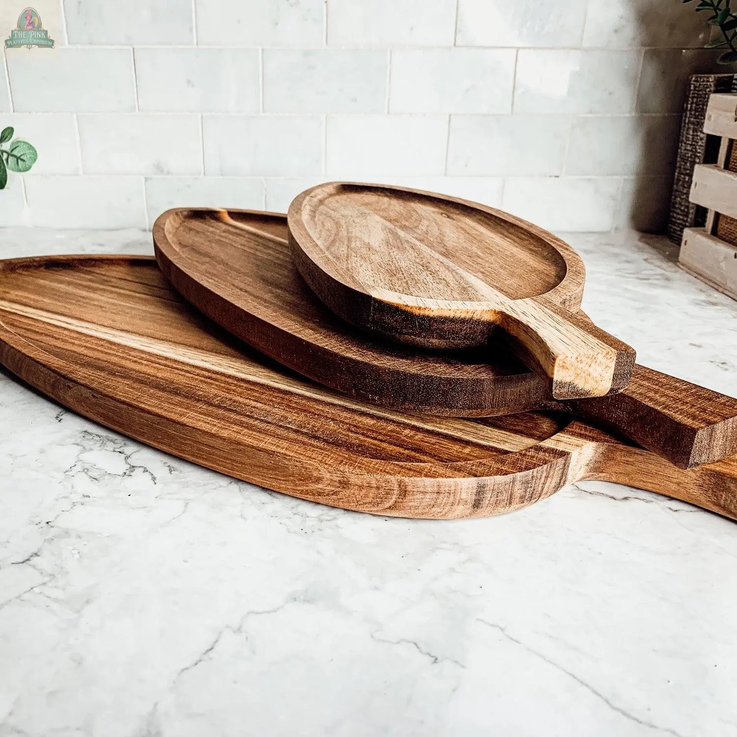 Three Nesting Trays in three sizes, crafted from acacia wood and featuring natural grain with paddle-shaped handles, are stacked on a white marble countertop against a tiled backsplash, offering an elegant serving solution.