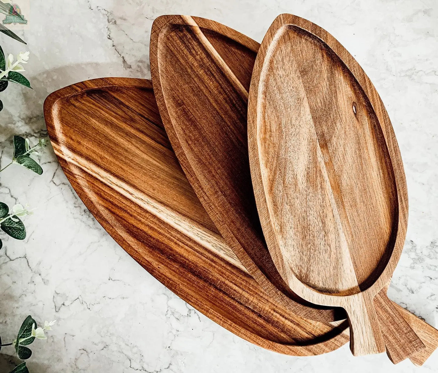 Three Nesting Trays in 3 sizes, crafted from acacia wood to resemble natural leaves, overlap on a light marble surface with green foliage visible in the top left, adding organic charm to your serving display.