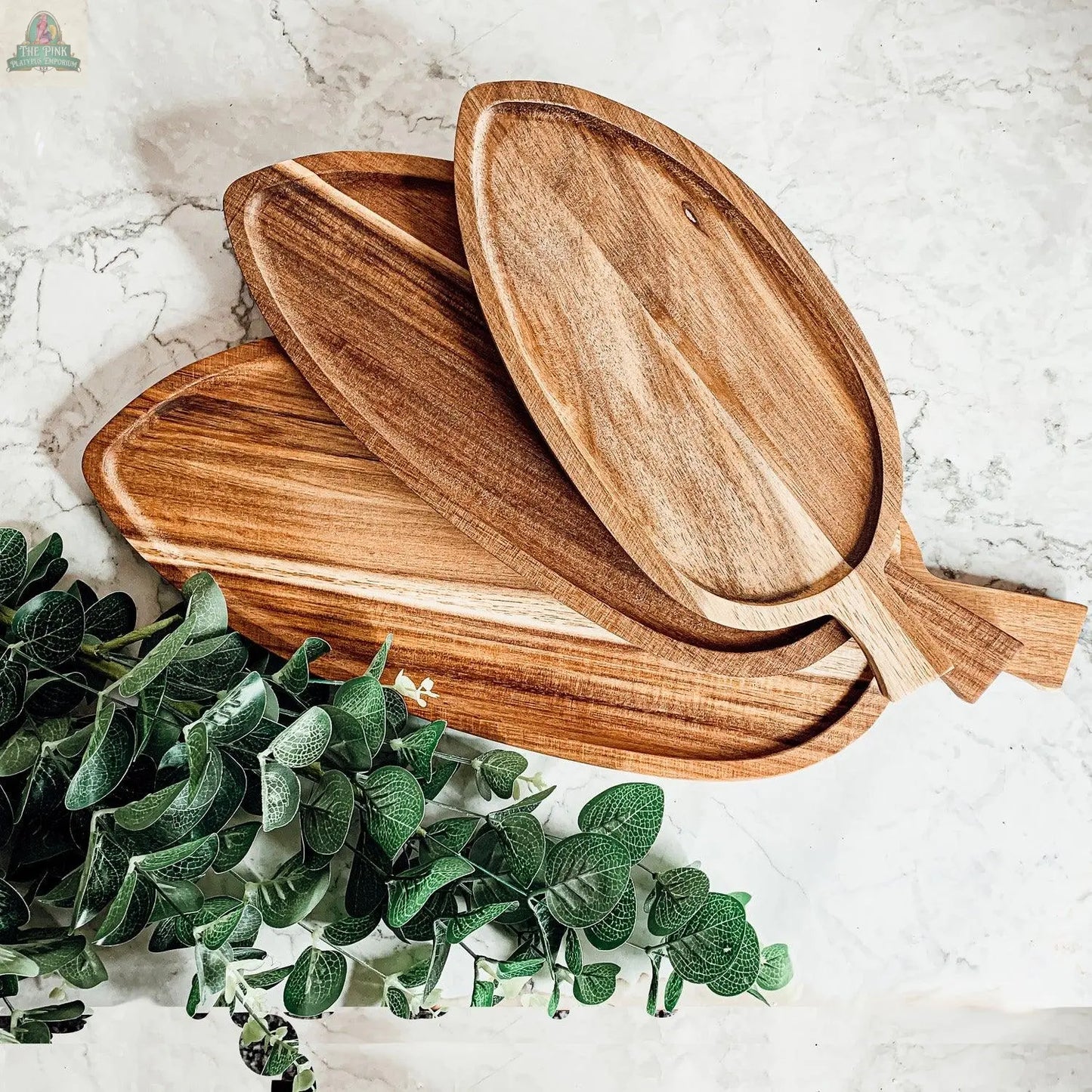 Three Nesting Trays in 3 sizes, made from acacia wood, are stacked on a white marble surface next to a green leafy plant.