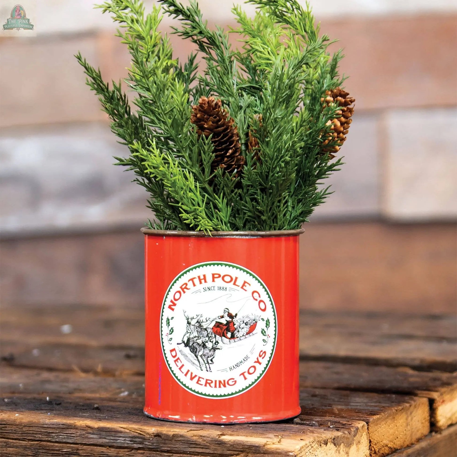 The North Pole Co Can displays artificial pine branches and pinecones, creating vintage holiday decor on a rustic wood surface with a soft-focus brick background.