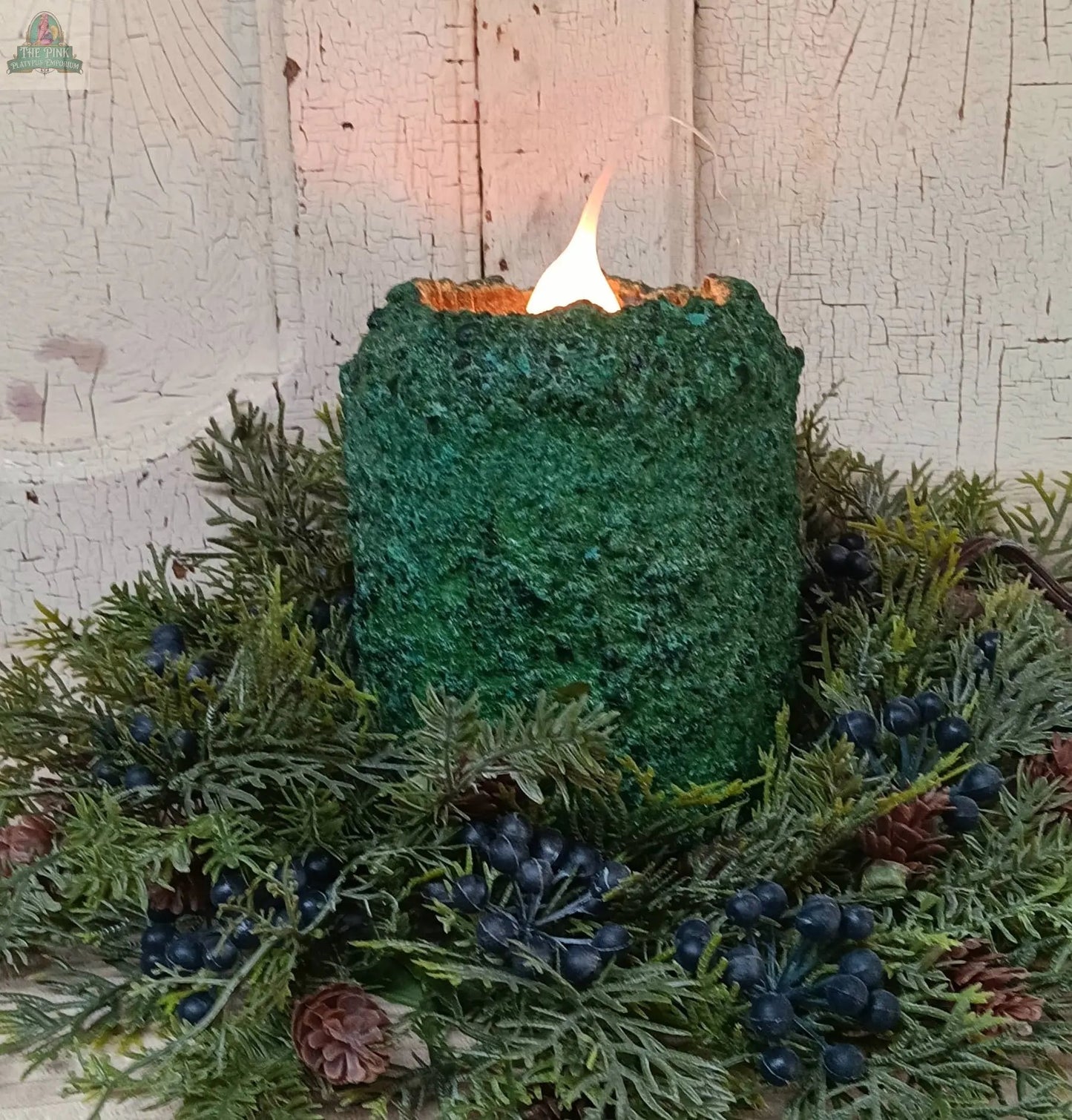 The Northern Pine Electric Candle, featuring a green textured finish, glows in the center of a wreath of evergreen branches, dark berries, and pinecones, set on a rustic, white, cracked wooden background.