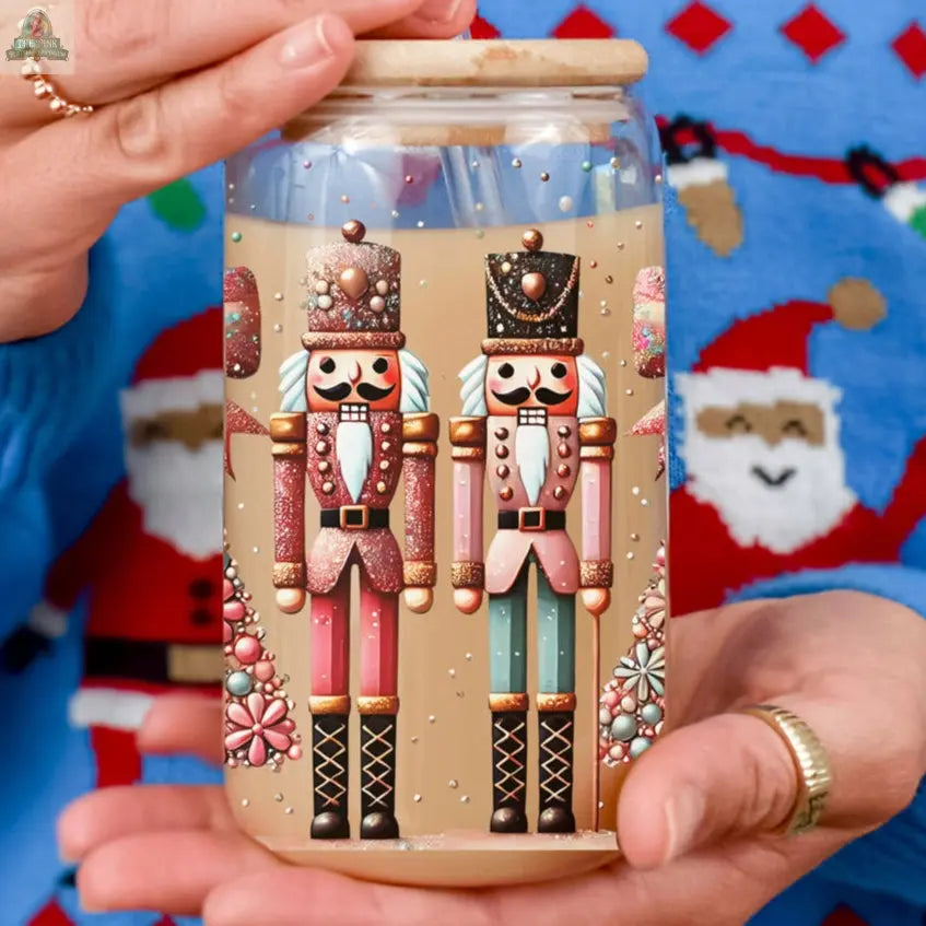 A person holds the Nutcracker Glam Soldier—a festive glass jar decorated with colorful nutcracker designs. Wearing a blue Christmas sweater with Santa faces and gold rings, they showcase this holiday drinkware filled with a light drink.