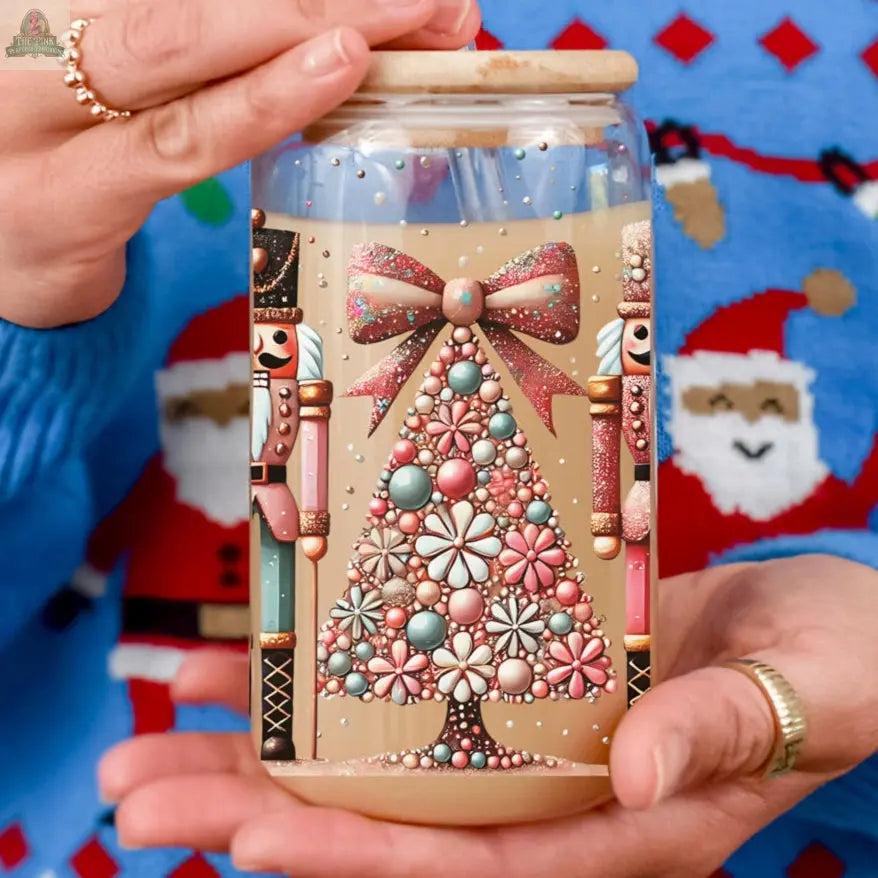 A person holds a decorated candle jar with a pastel floral Christmas tree and a bow, flanked by two Nutcracker Glam Soldier tumblers, while wearing a festive blue sweater with Santa faces.